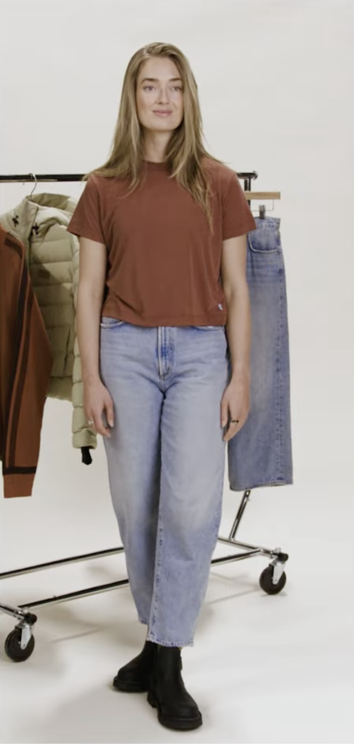 A young woman with long blonde hair standing in a clothing studio, wearing a rust-colored t-shirt, light blue jeans, and black boots, with clothing hanging on a rack behind her.