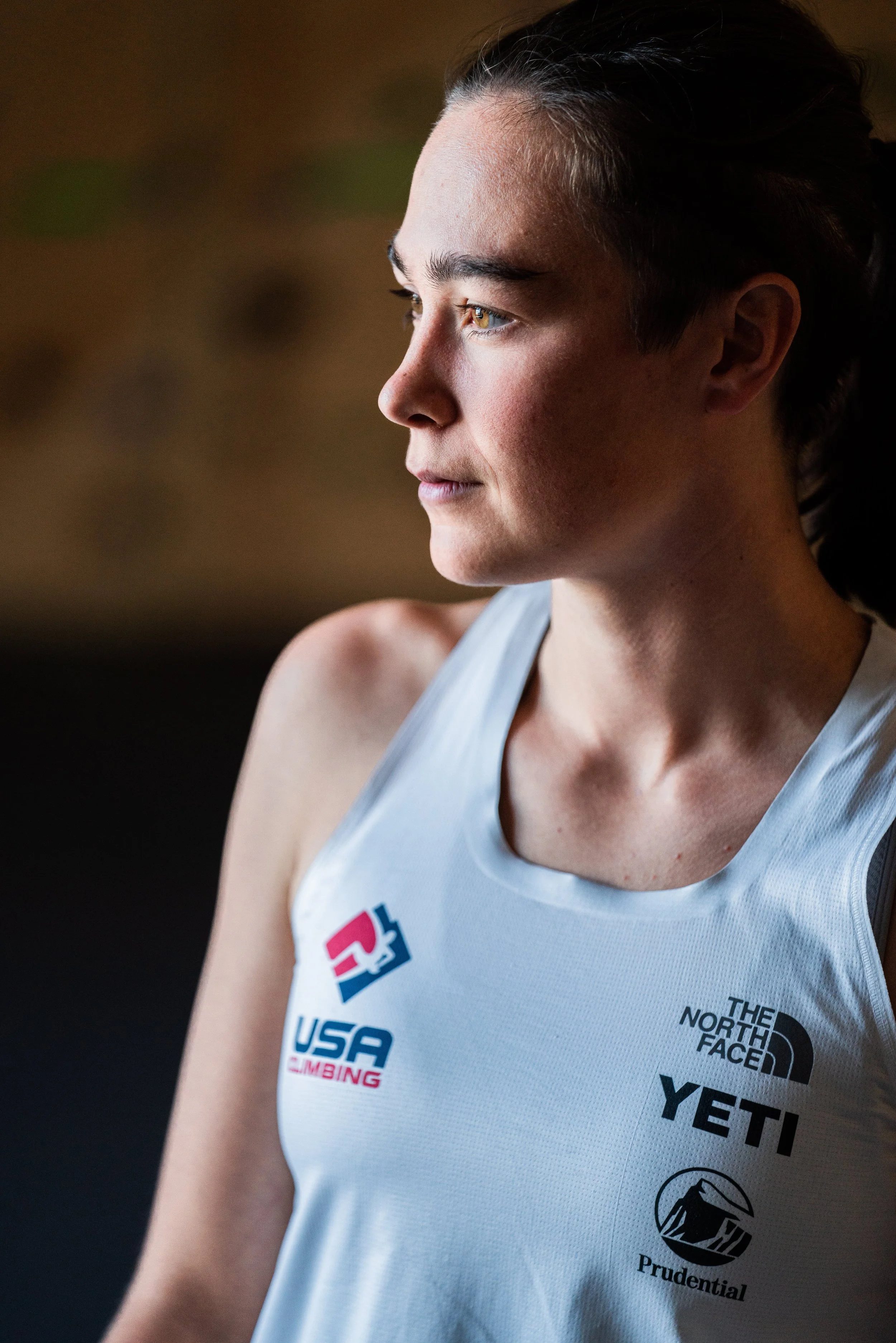 Close-up side view of a young woman with dark hair in a ponytail, wearing a white sleeveless sports jersey with various sponsor logos, looking out thoughtfully.