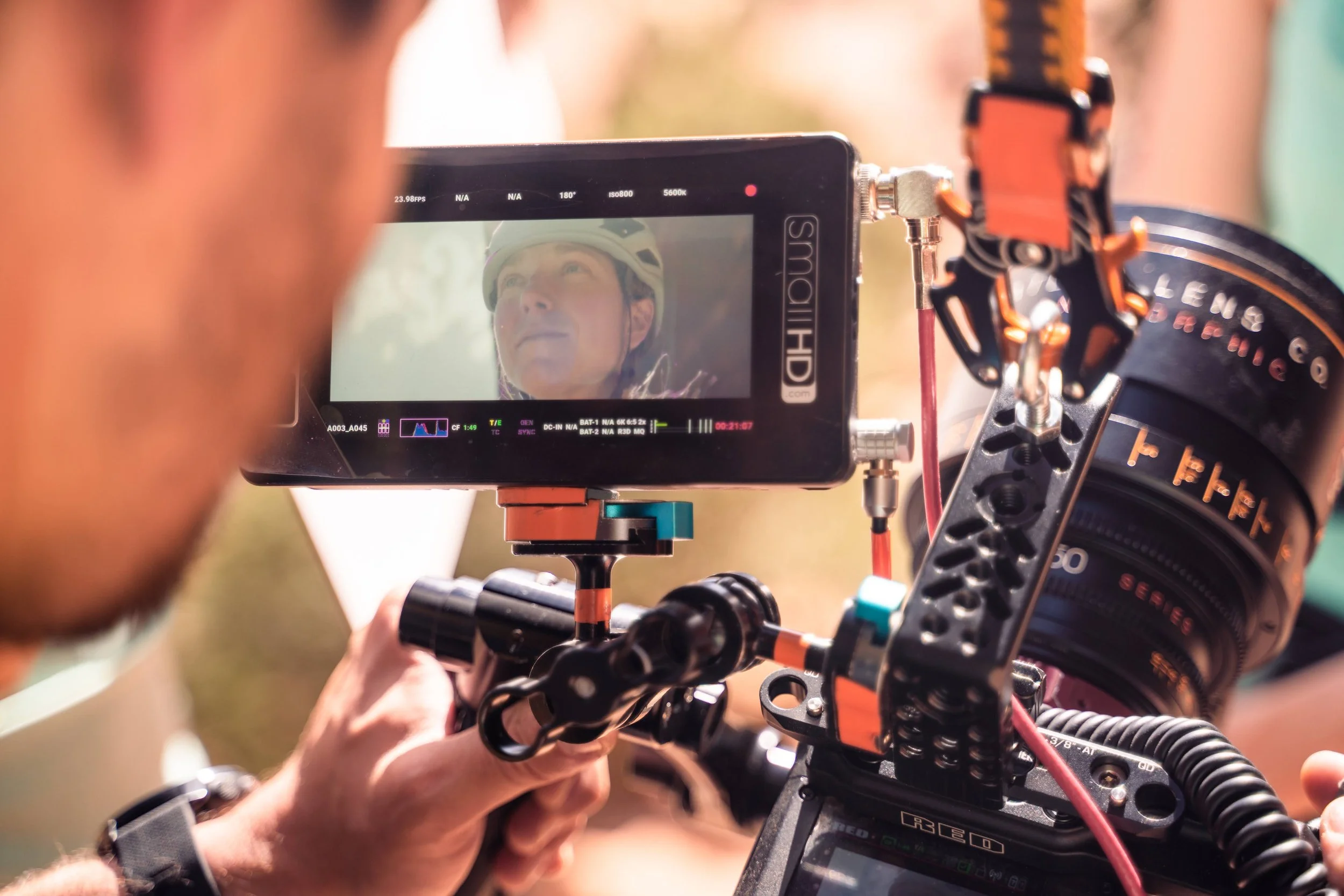 A person operating a professional camera monitor mounted on a camera rig, with the person's face and a helmet reflected on the monitor screen.
