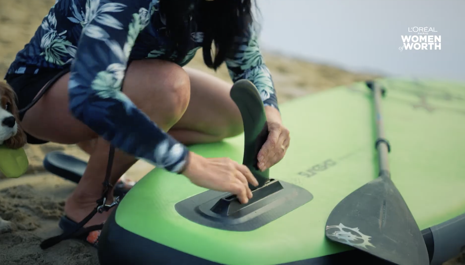 A person in a floral shirt is kneeling on a sandy beach, attaching a fin to a lime-green paddleboard, with a dog lying beside them.