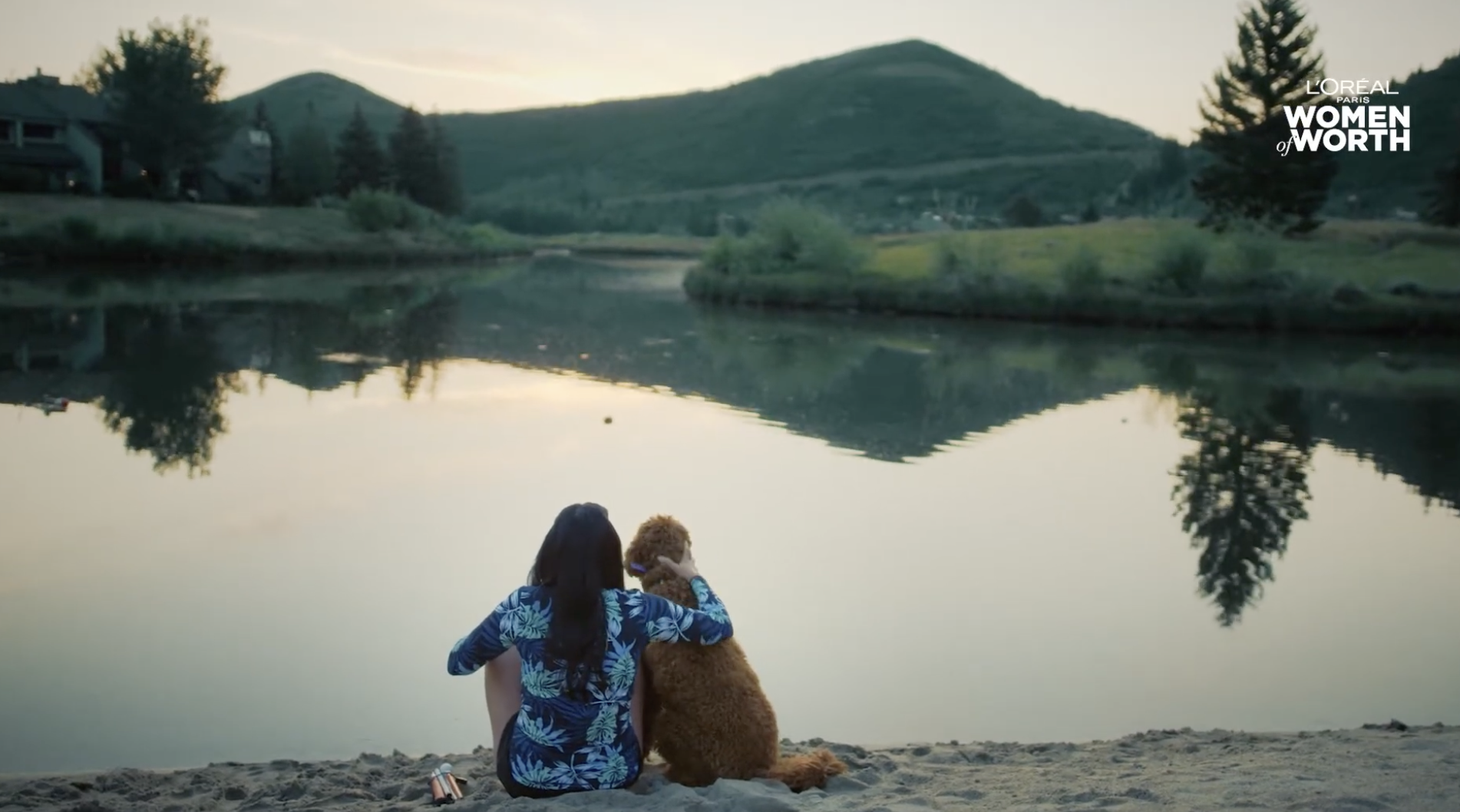 A person with long dark hair, wearing a blue floral shirt, sitting on a sandy beach with a brown dog, looking out over a calm river surrounded by green hills and trees, during sunset or dawn.