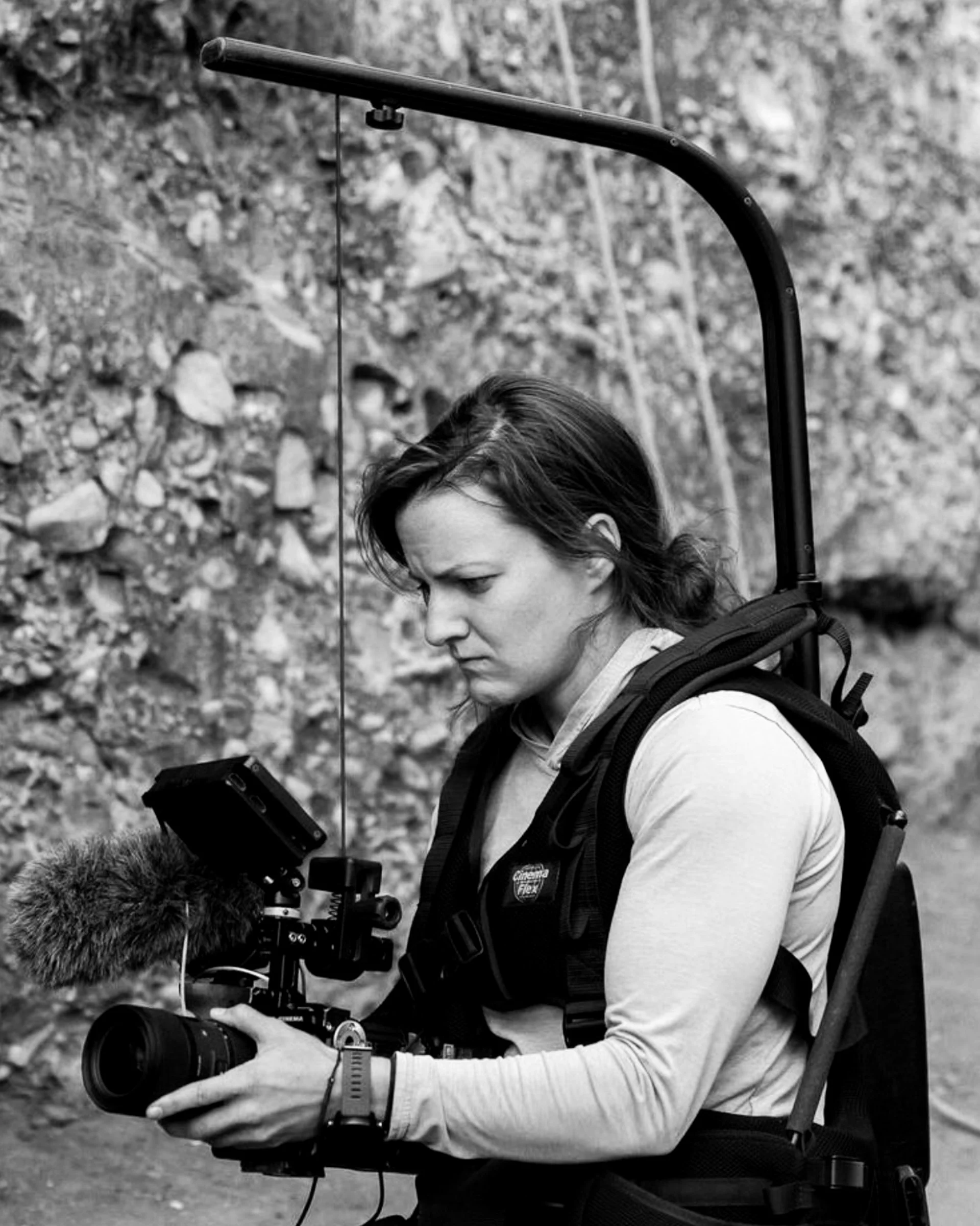 Person with long hair, wearing a backpack, operating a professional video camera outdoors with a rocky background.