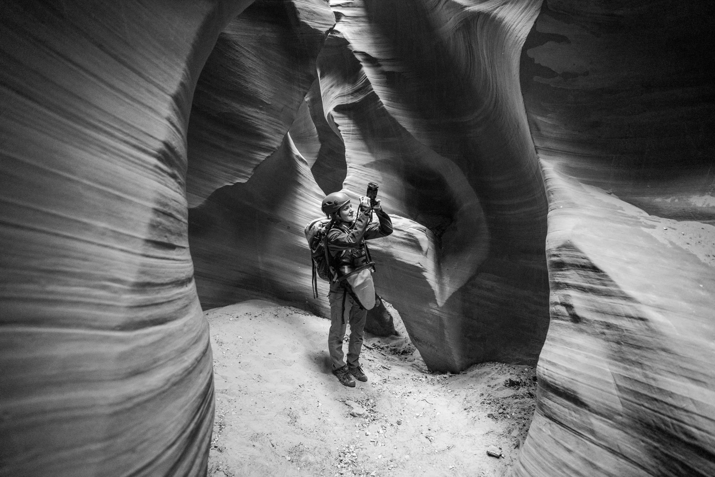 Person wearing a helmet and backpack, taking a photo with a smartphone inside a narrow slot canyon with smooth, curved sandstone walls.
