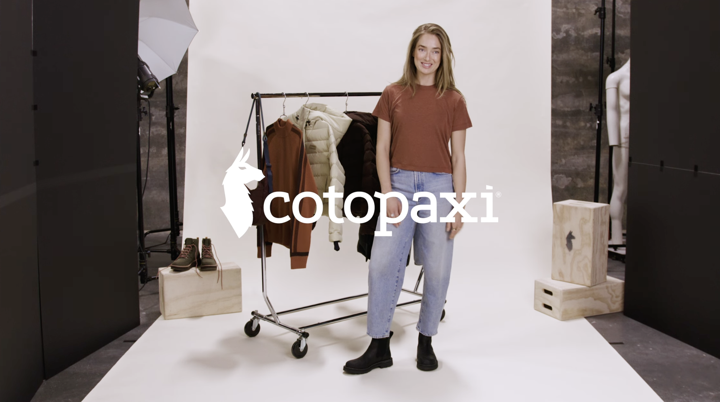 A woman standing next to a clothing rack with jackets and shirts, in a photo studio with lights and backdrop, featuring the Cotopaxi logo in the foreground.