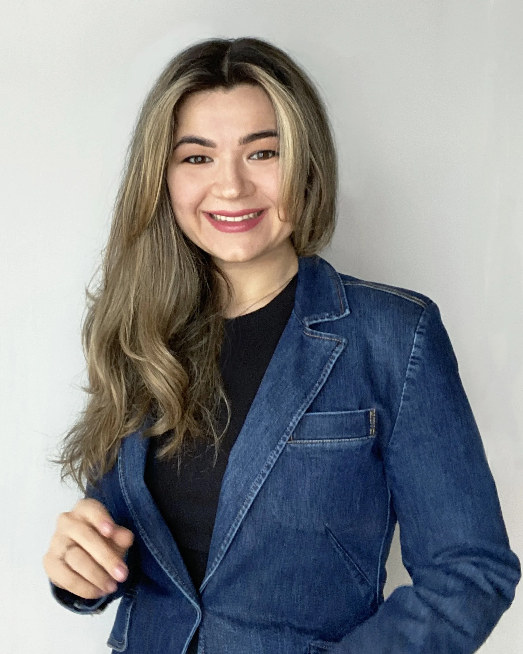 A woman with long, wavy blonde hair smiling, wearing a black top and a blue denim jacket, standing against a plain white background.