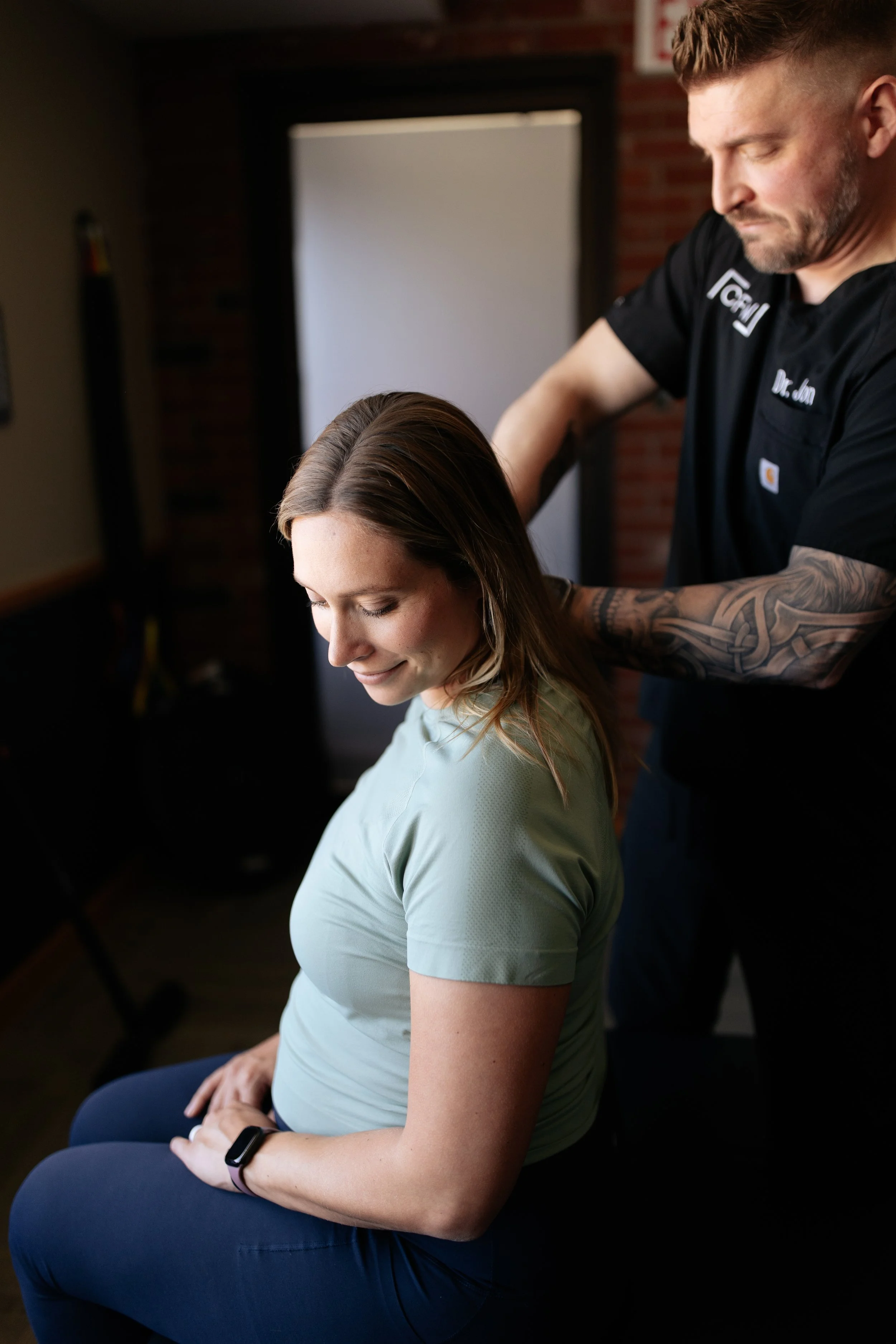 A dentist or medical professional with tattoos on their arm attending to a woman with brown hair, wearing a light green shirt, in a setting with brick walls and a dark background.