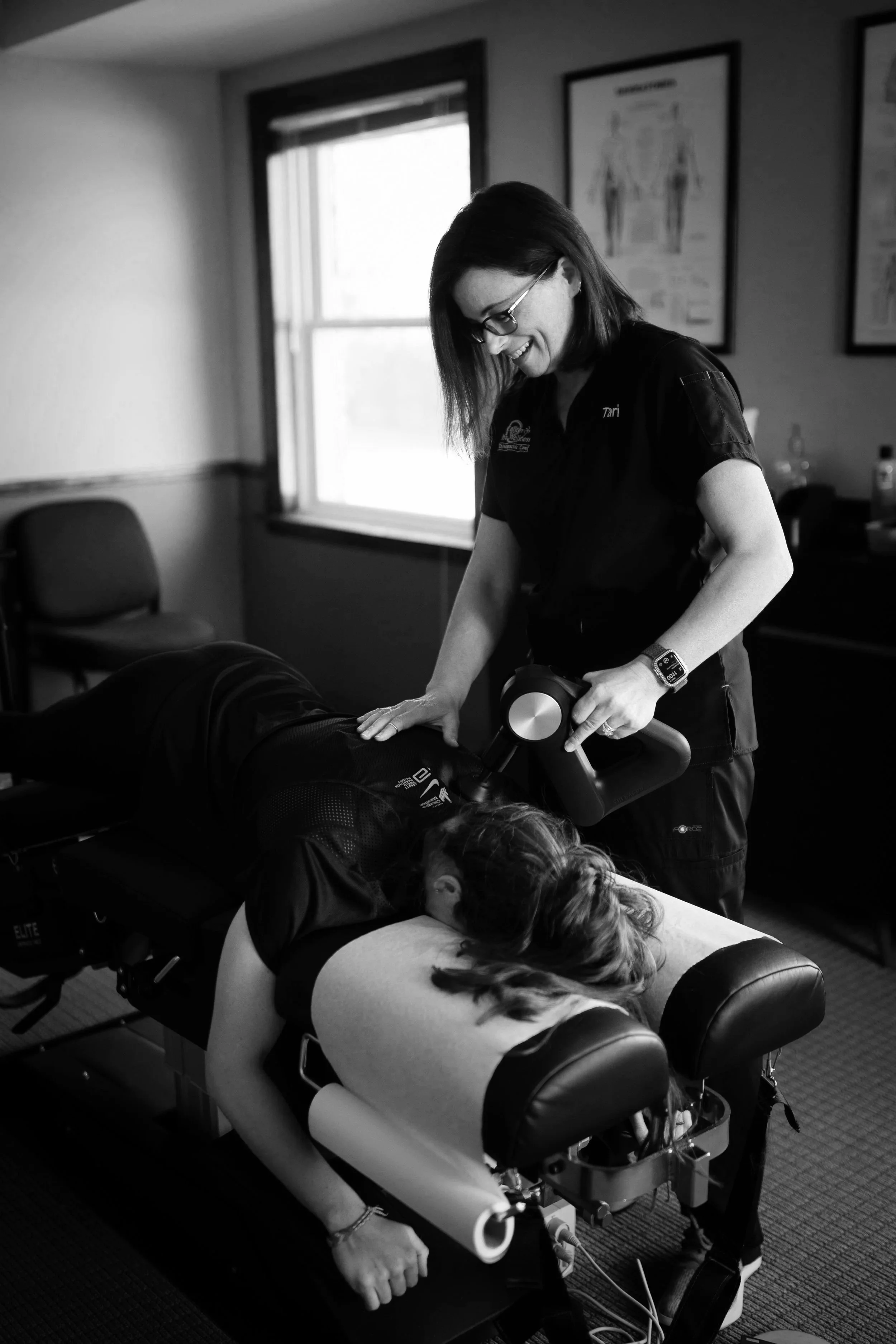 A woman receiving physical therapy from a therapist on a massage table. The woman is lying face down with her head turned to the side, and the therapist is standing next to her, holding a massage device. The room has a window with blinds and framed educational posters on the wall.
