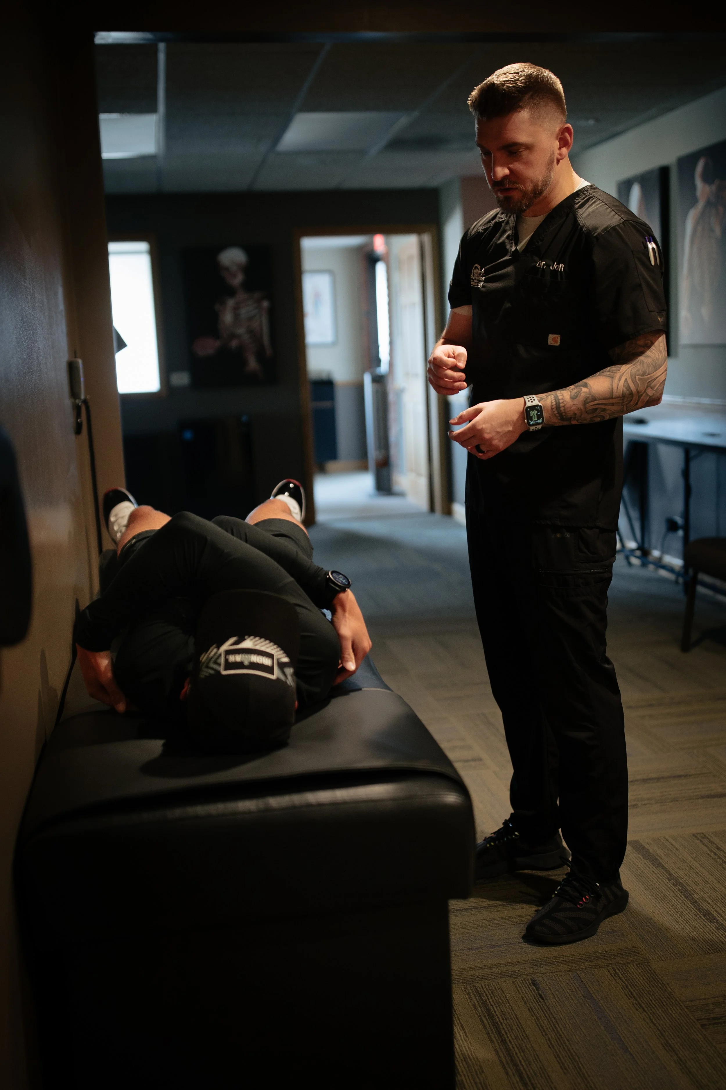 A man in black medical scrubs talking to a person lying on a black leather examination table in a medical setting.
