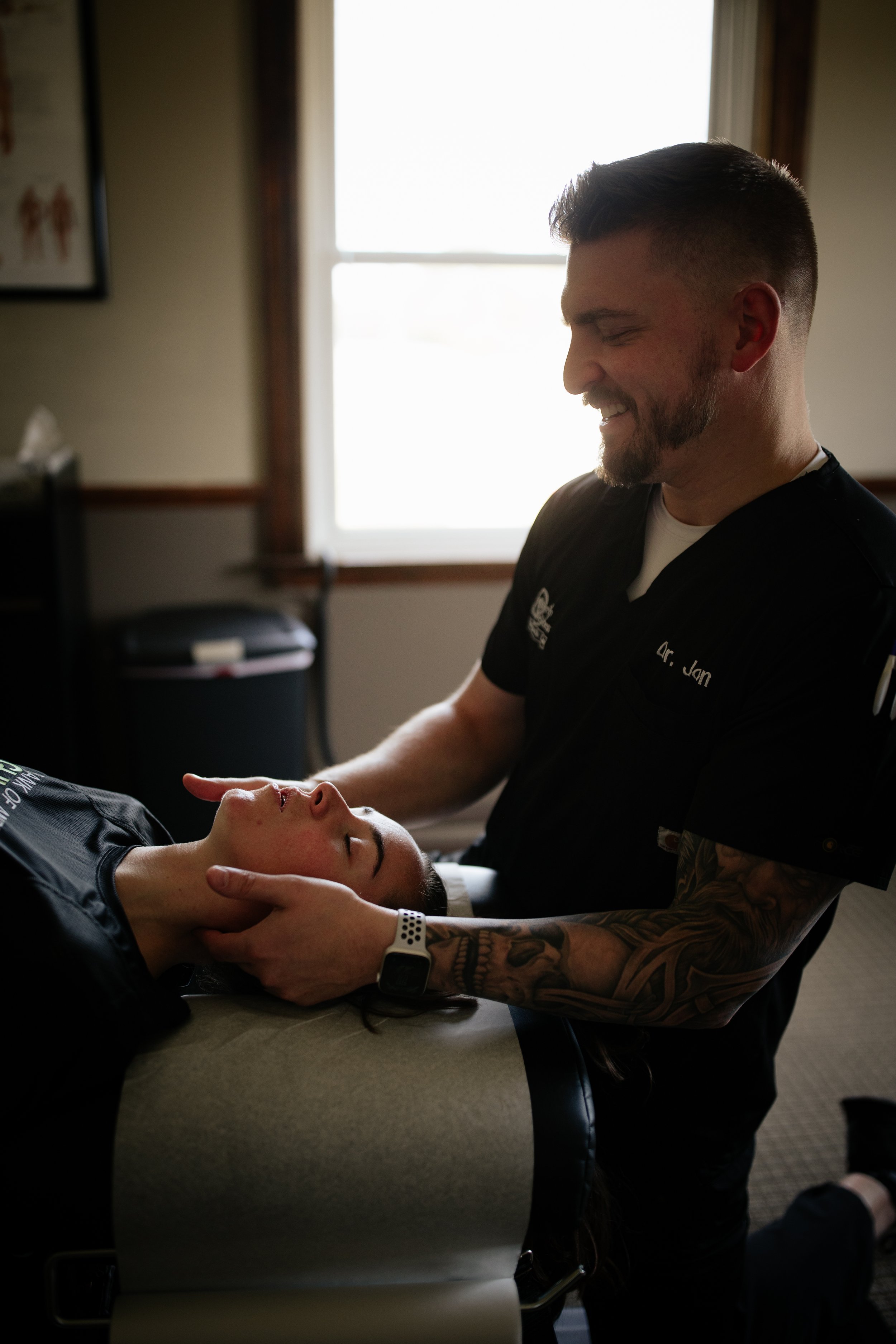 A man in medical scrubs smiling while looking at a woman lying on a treatment table during a chiropractic or massage therapy session.