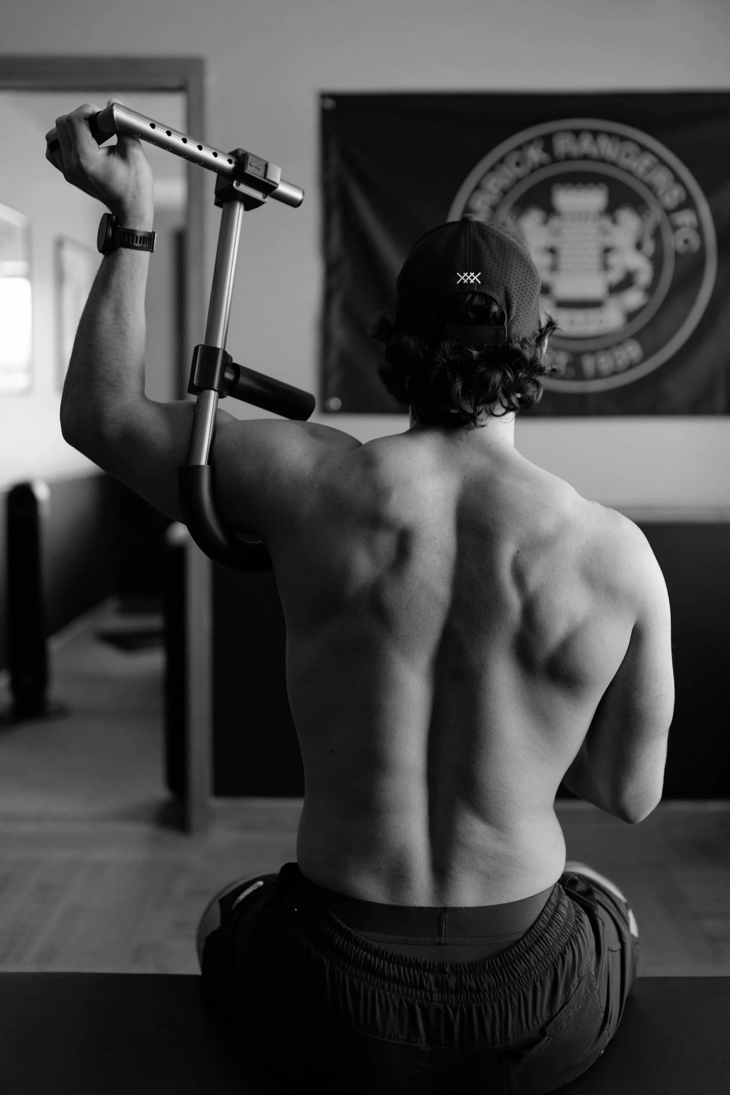 A shirtless man with curly hair, wearing a cap backwards, seated on a yoga mat, lifting a crutch in a room with a flag or banner in the background.