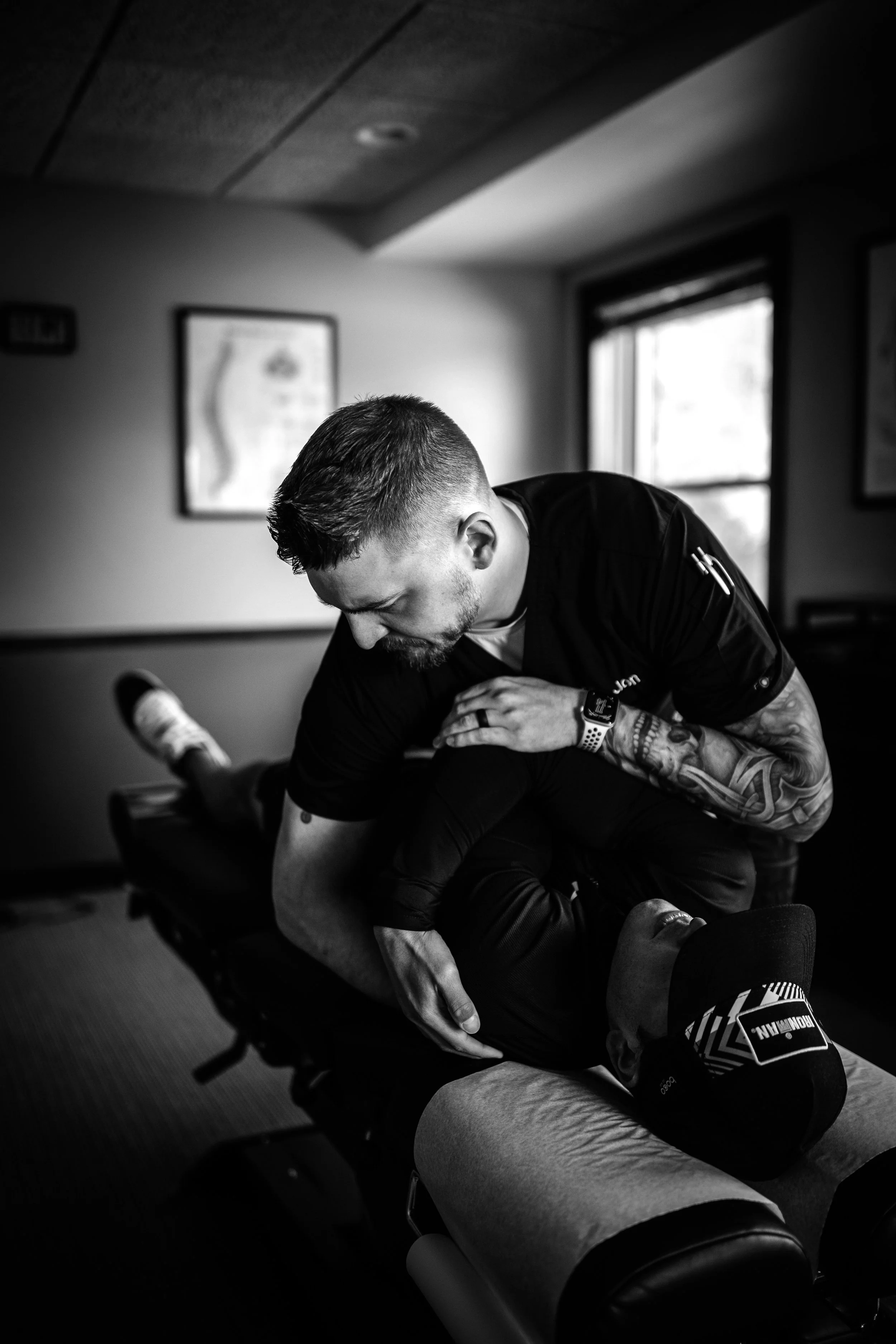 A chiropractor or physical therapist performing a treatment on a patient lying face down on a treatment table in a room with framed artwork and a window.