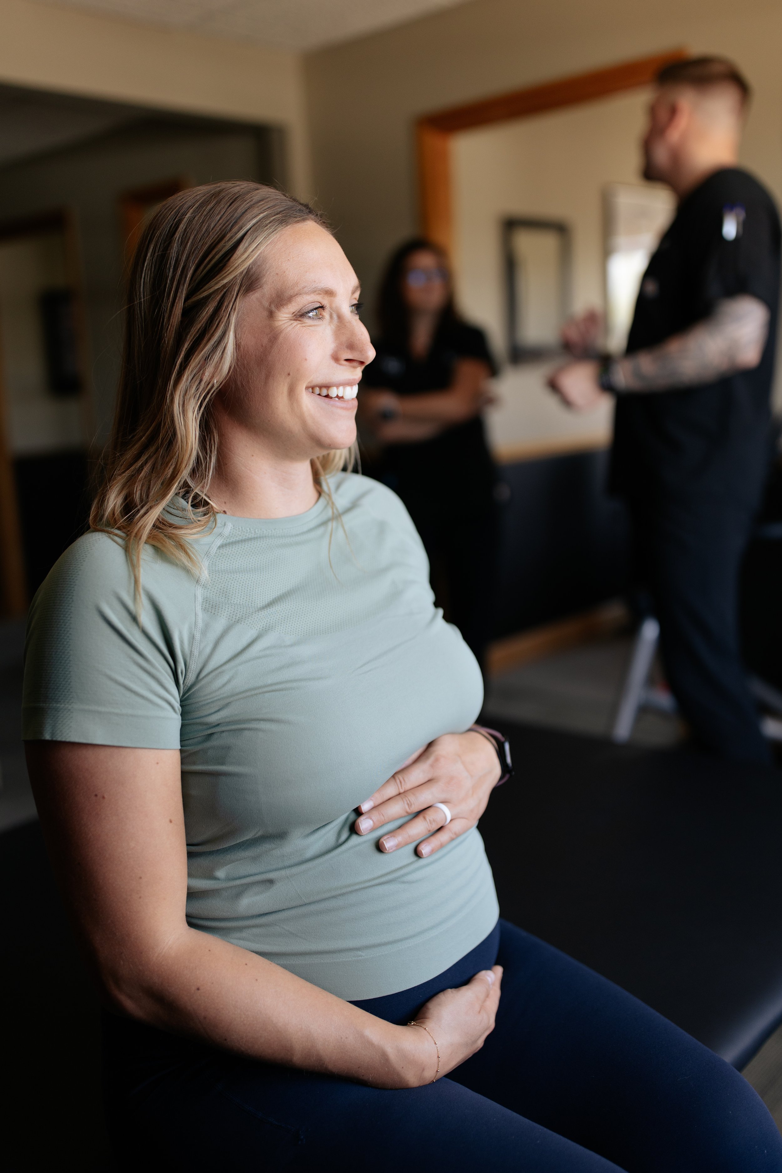 Pregnant woman sitting with hands on her belly, smiling, in a room with blurred background including a woman and a man standing.