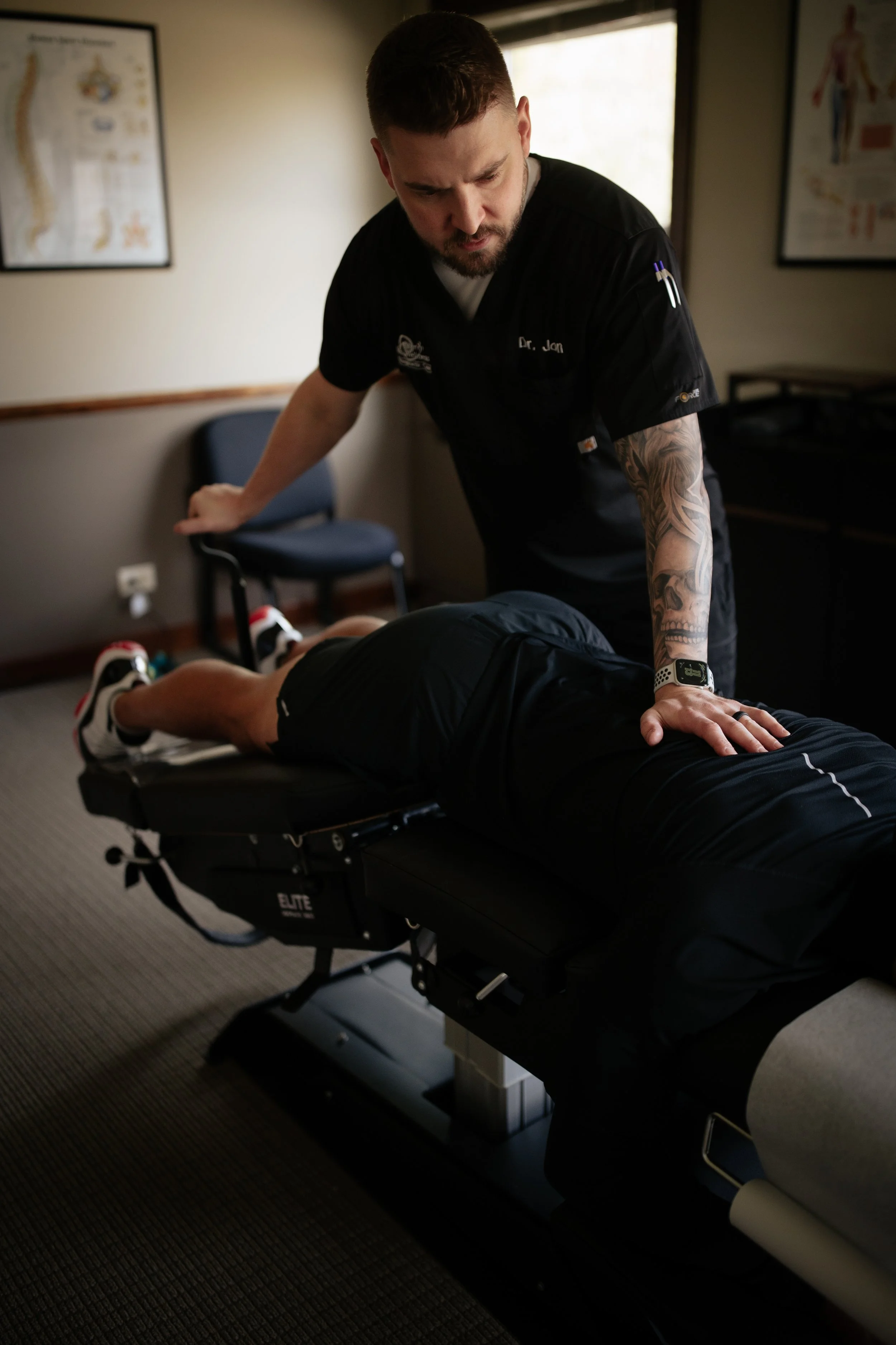A healthcare professional performs a physical therapy session on a patient lying face down on a medical examination table in a room with medical posters on the wall.