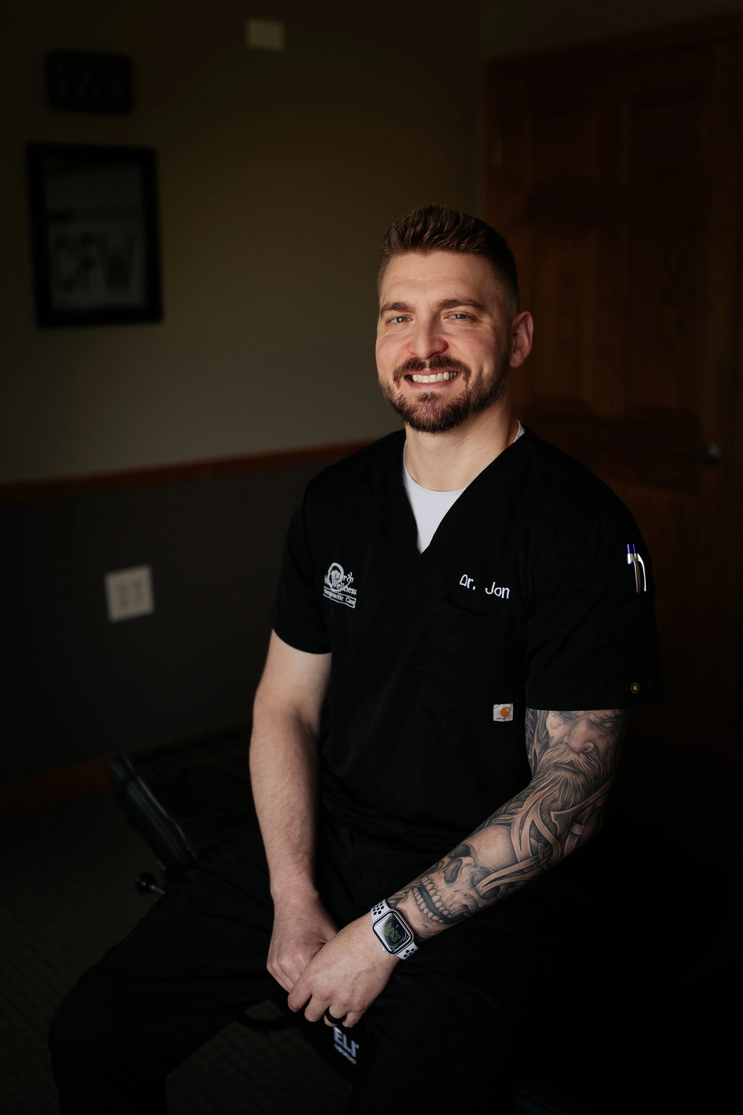 A smiling man with a beard, wearing a black medical uniform with 'Dr. Jon' embroidered, sitting indoors on a black bench. He has a large tattoo sleeve on his right arm and wears a smartwatch. The background includes a wooden door, framed picture, and electrical outlets.