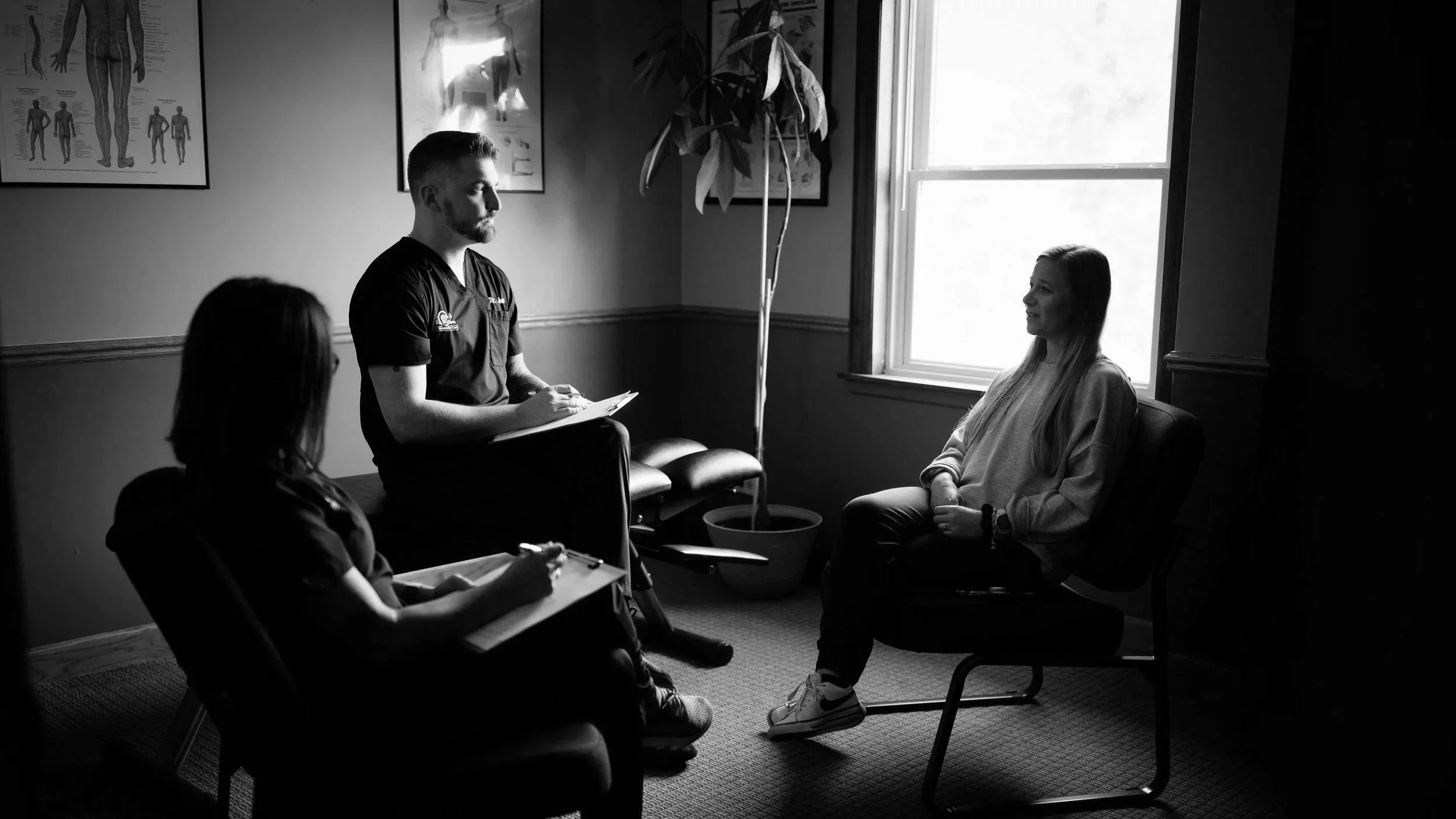A black and white photo of a medical consultation with a healthcare professional and three women. The doctor stands near a window, holding a notepad, talking to a seated woman with long blonde hair. Two women are seated in the foreground, one holding a clipboard and pen, and the other with her back turned, listening attentively.