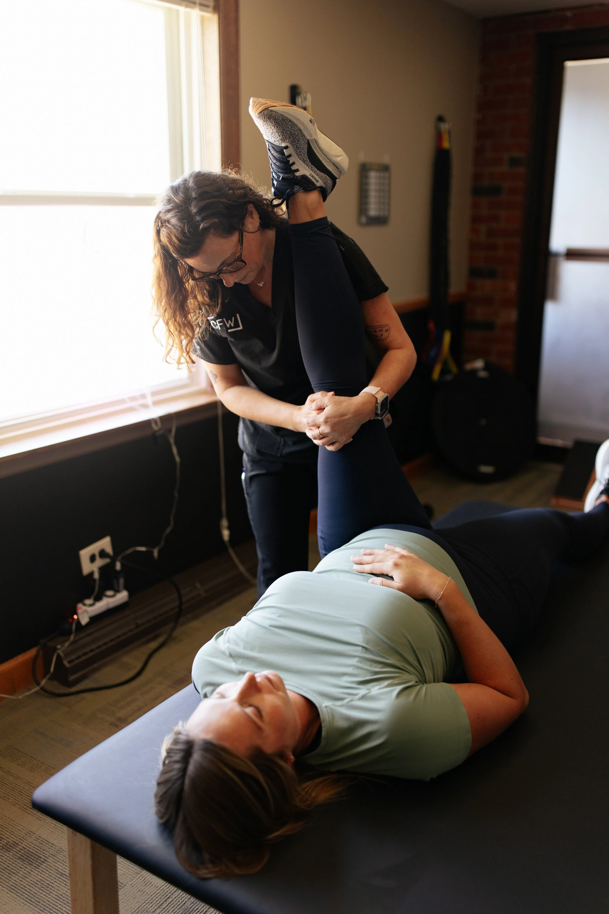 A woman assisting a person with a physical therapy exercise on a massage table in a well-lit room.