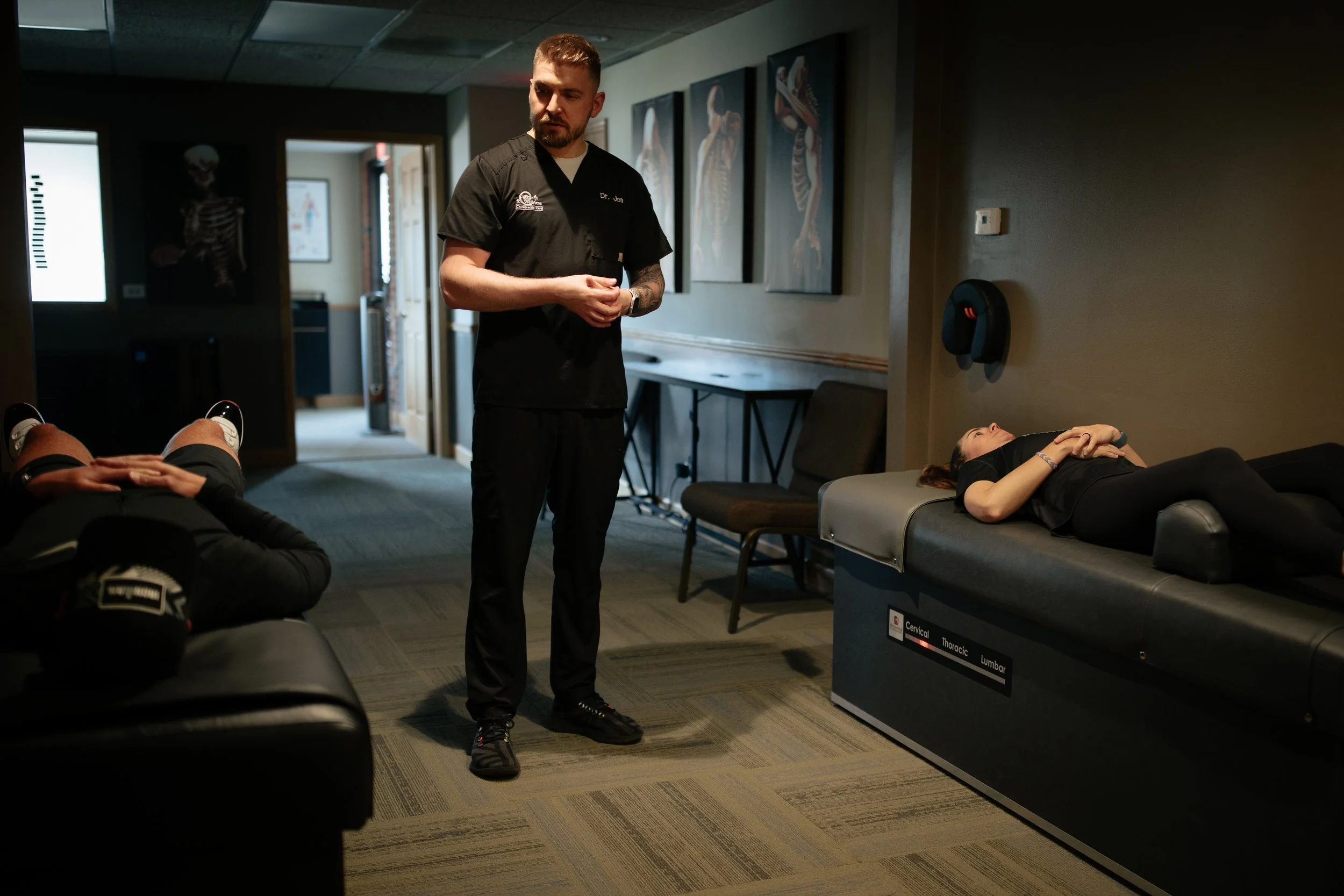 A healthcare professional standing in a medical clinic or physical therapy office, with two patients lying on treatment tables, while skeletal anatomy photographs are displayed on the wall behind him.