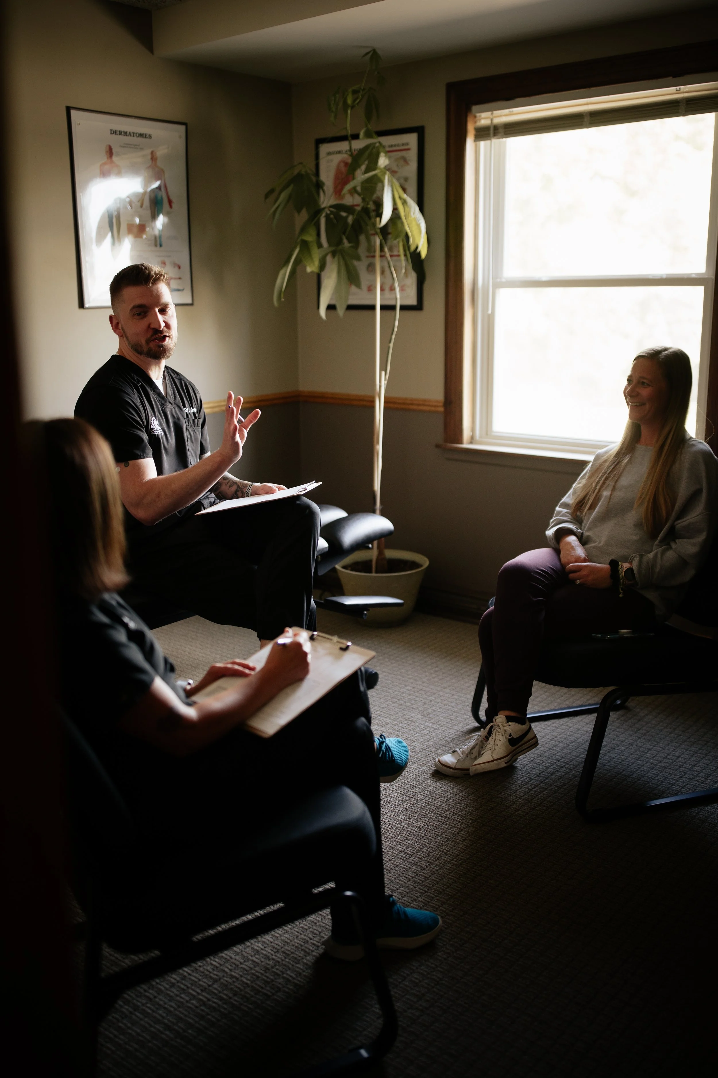 Medical professional conducting a consultation with a woman in a small office, with another person taking notes, and a large window letting in natural light.