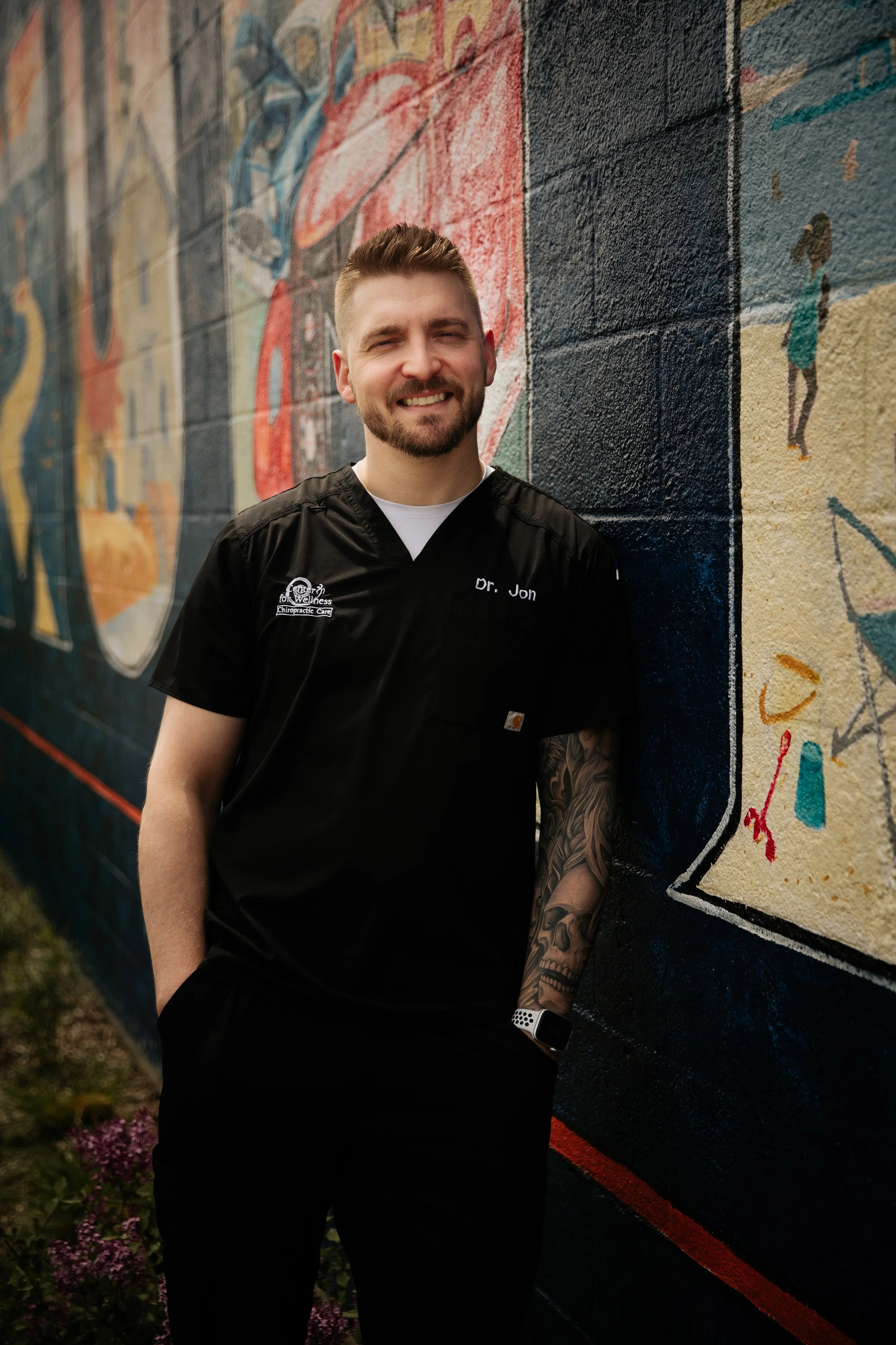A smiling man in a black medical uniform with the name tag Dr. Jon is leaning against a colorful mural painted on a brick wall.