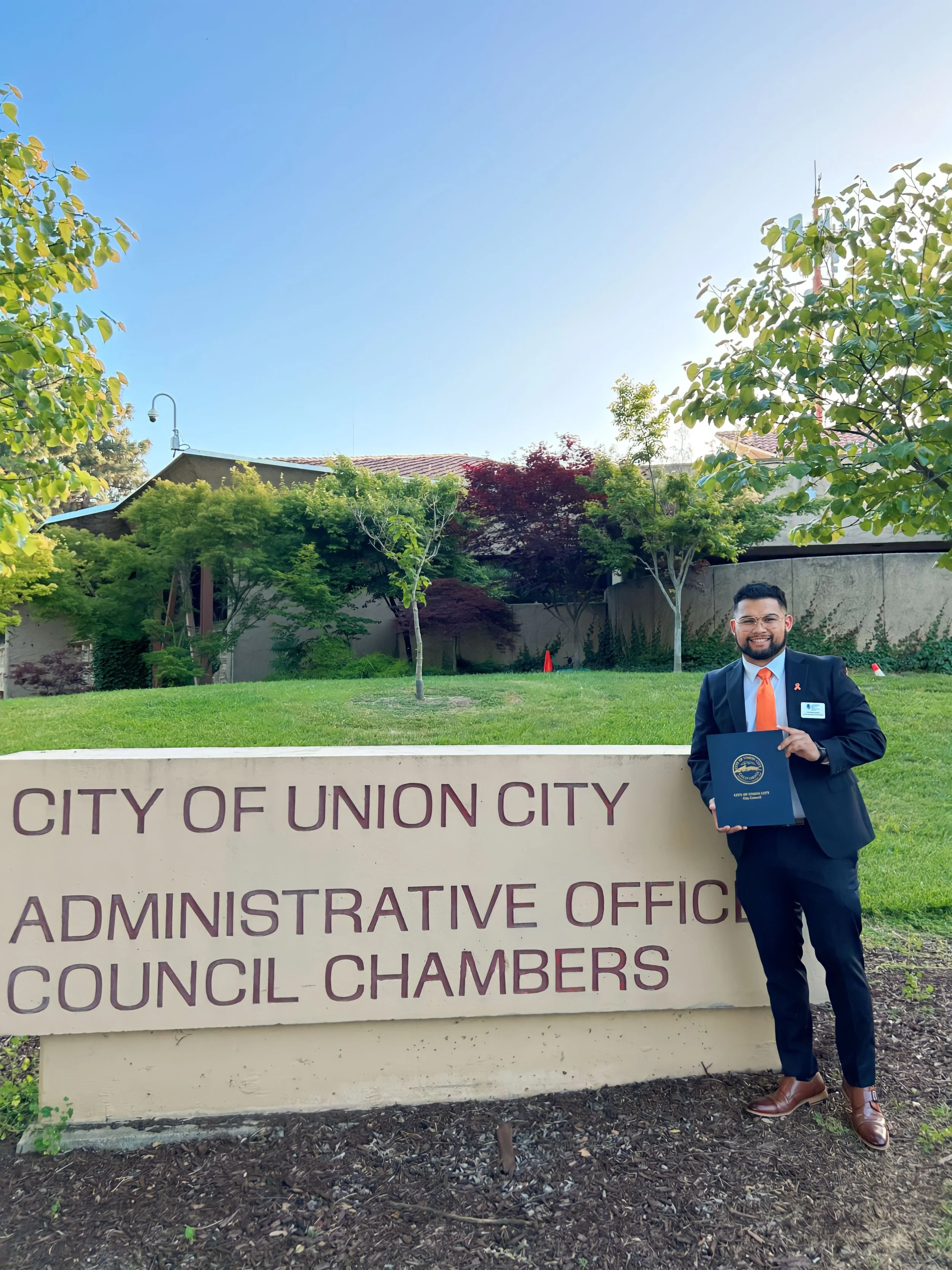 A man in a suit holding a folder standing next to a sign that reads 'City of Union City Administrative Office Council Chambers' outside a government building with trees and a grassy area.