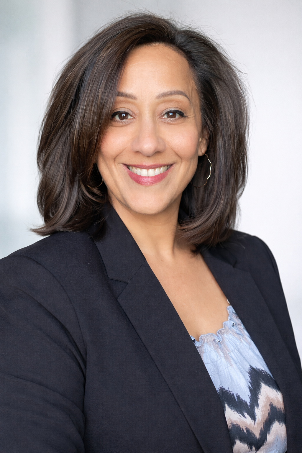 A professional woman smiling, wearing a black blazer and a patterned top, with shoulder-length brown hair and hoop earrings.