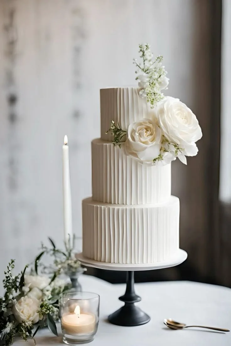 A three-tier white wedding cake with vertical textured piping, decorated with white roses and greenery, on a black cake stand with candles and floral arrangements nearby.