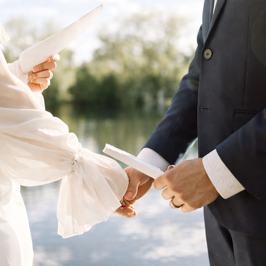 A couple holding hands during a wedding ceremony outdoors near a body of water, exchanging rings.
