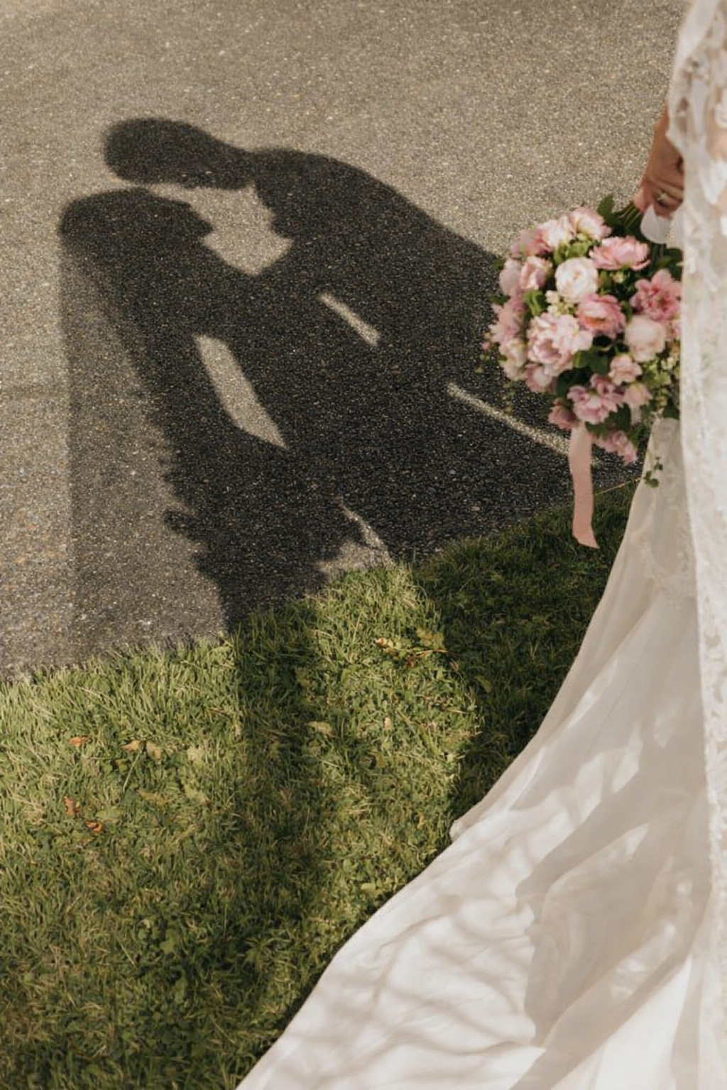 Shadow of a bride holding a bouquet of pink flowers, with the shadow of a groom on the ground, cast on pavement beside a grassy area.