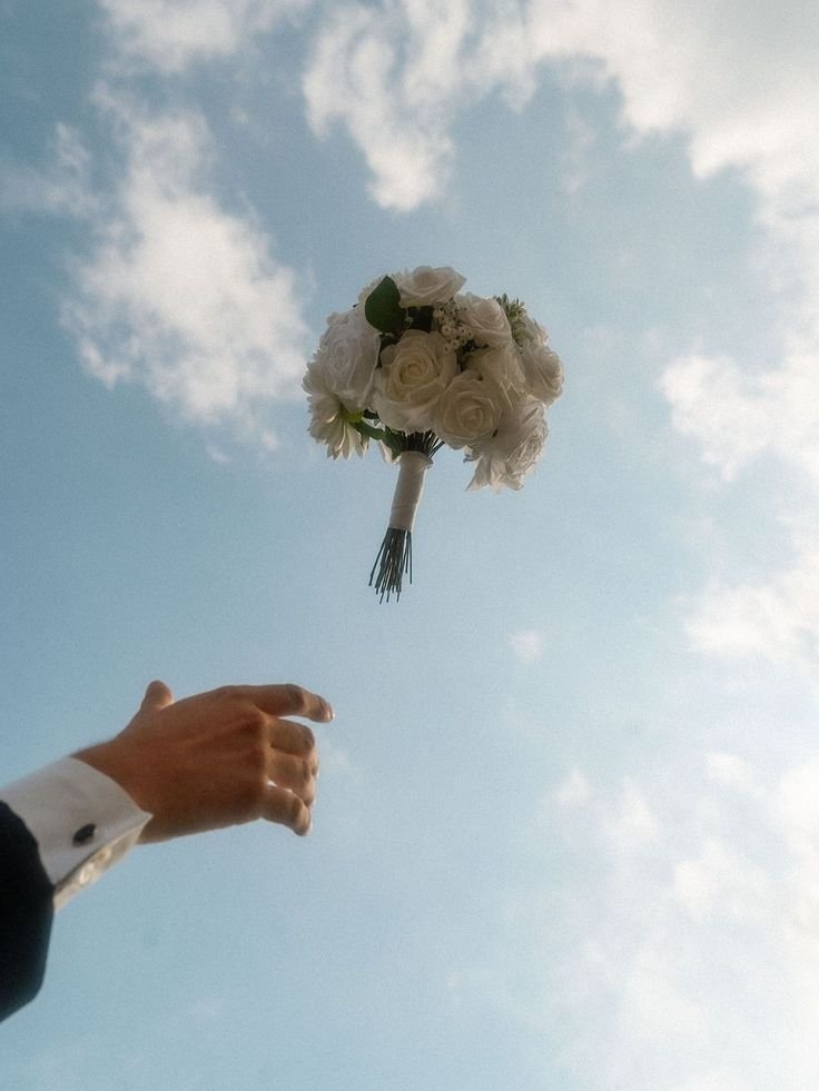 A hand in formal attire releasing a bouquet of white roses into the sky against a backdrop of blue sky and scattered clouds.