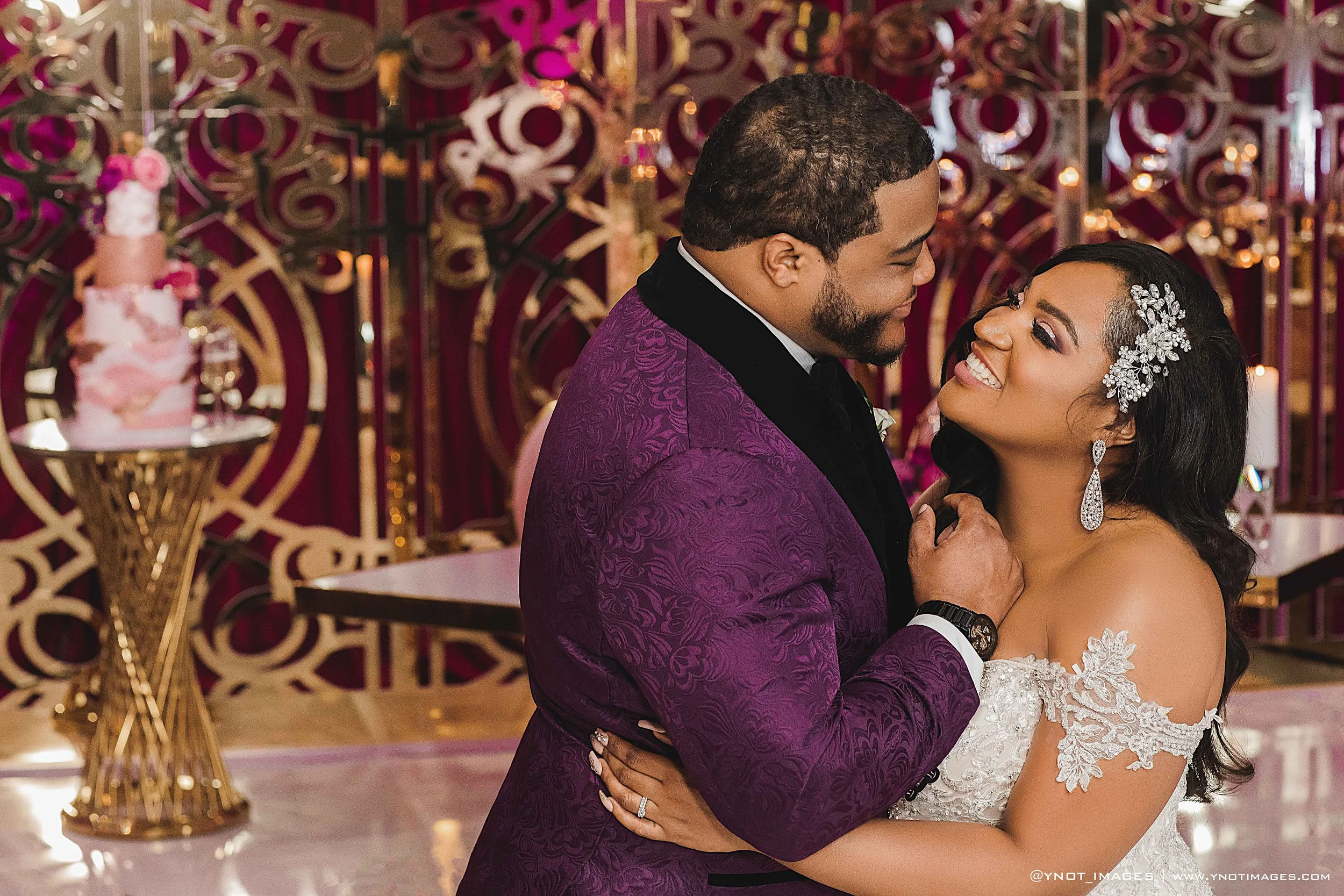 A newlywed couple shares a dance at their wedding reception. The groom is dressed in a purple tuxedo with a black collar, and the bride is in a white lace wedding gown with floral embellishments and decorative hairpiece. They are smiling and looking at each other, with a decorative pink and gold wedding cake and ornate background in a banquet hall.