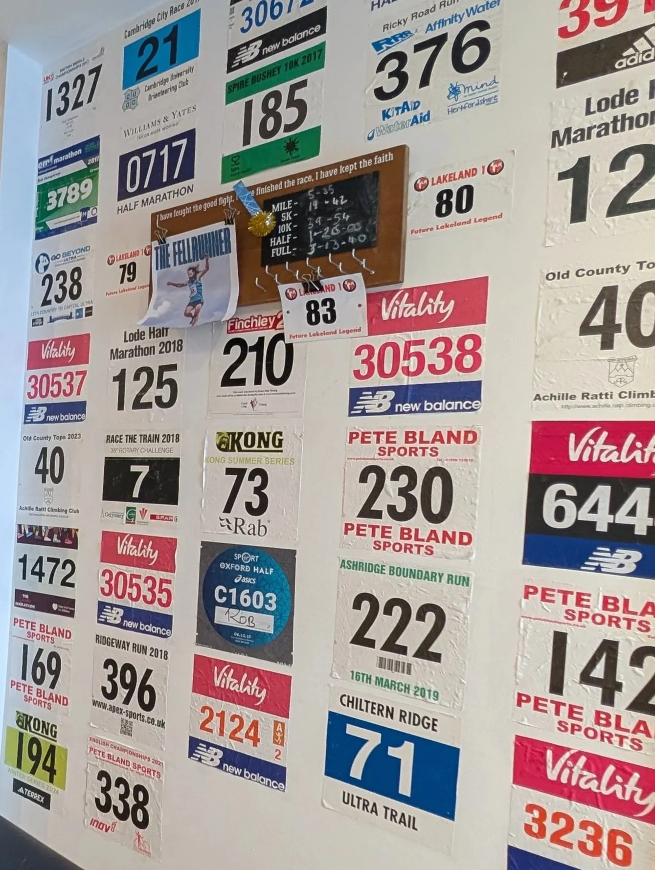 A wall covered with various marathon race bibs, sports event stickers, and a small chalkboard with race times. The bibs are in different colors and sizes, each containing numbers and event details.