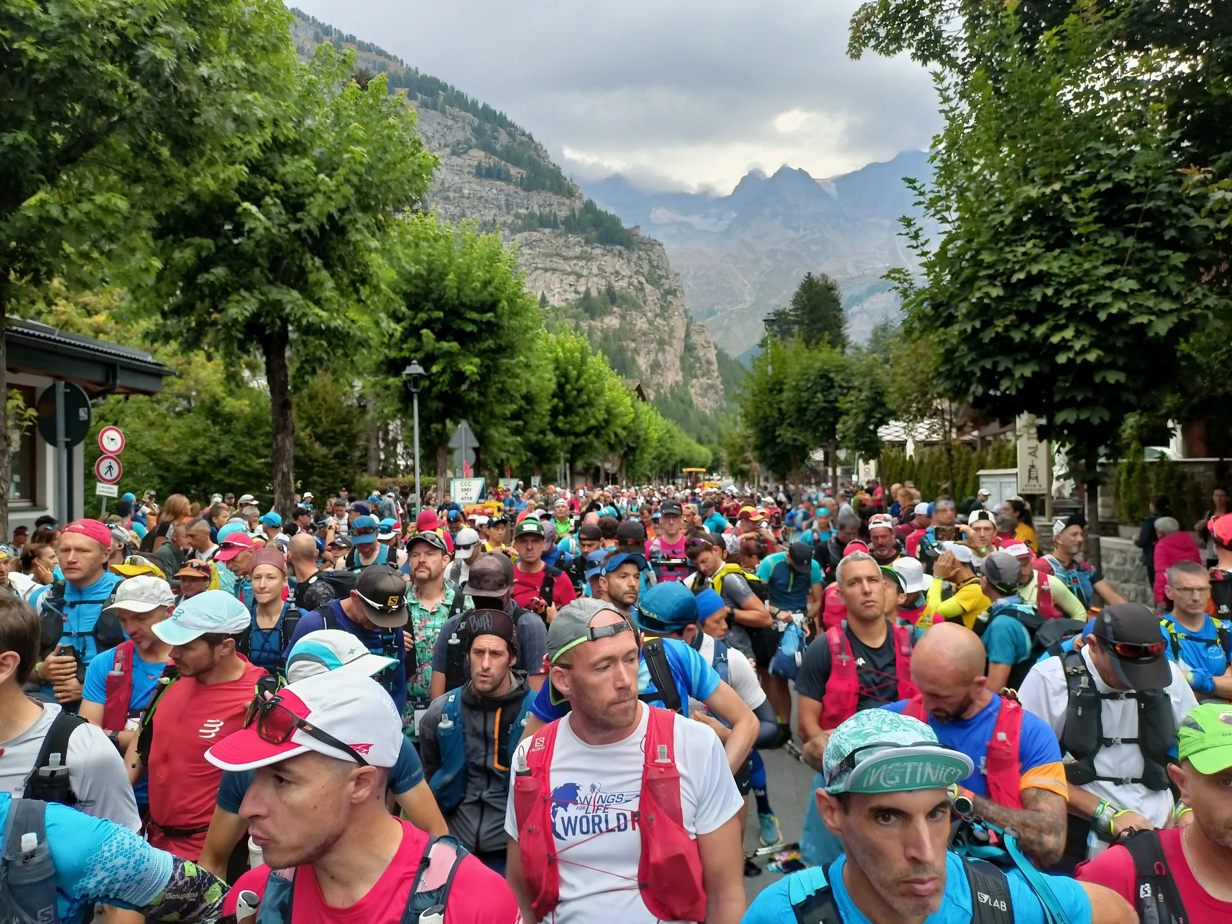 A large group of marathon runners gathered on a tree-lined street with mountains in the background, preparing for the start of the UTMB race.