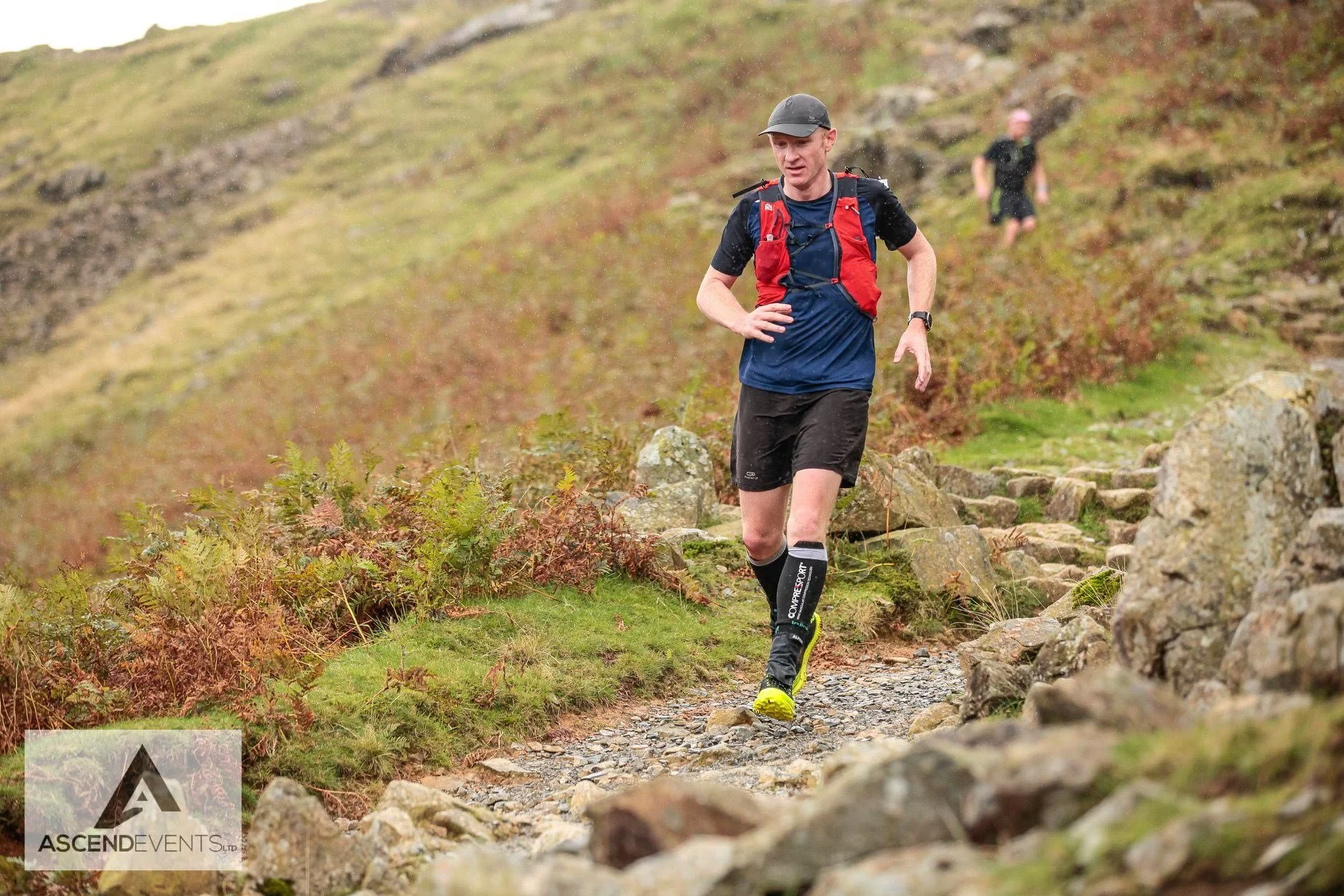 A male trail runner in black shorts and a blue shirt with a red hydration vest runs along a rocky mountain trail with grass and shrubs. There is another runner in the background along the same path.