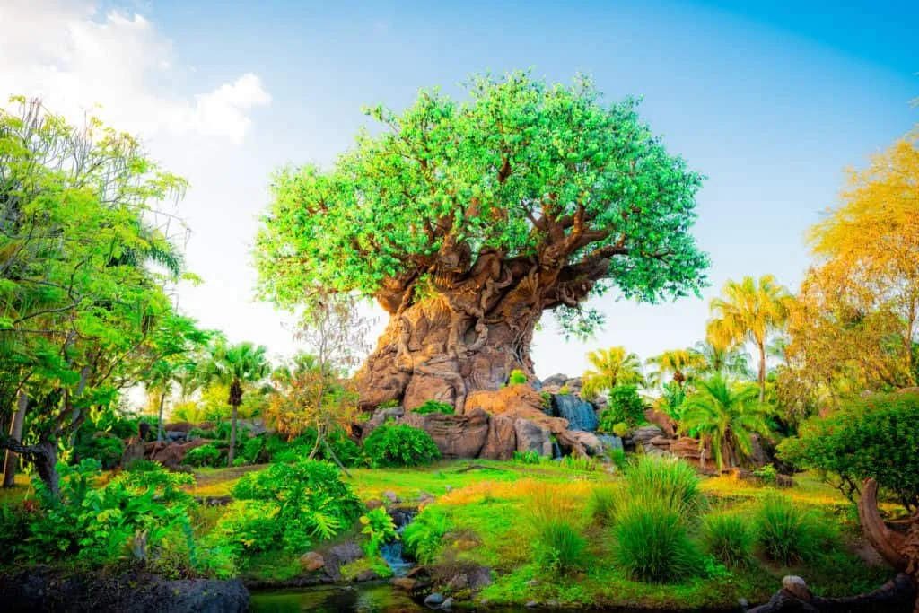 A large artificial tree with a thick trunk and lush green leaves, surrounded by tropical plants and small waterfalls in a landscaped garden.