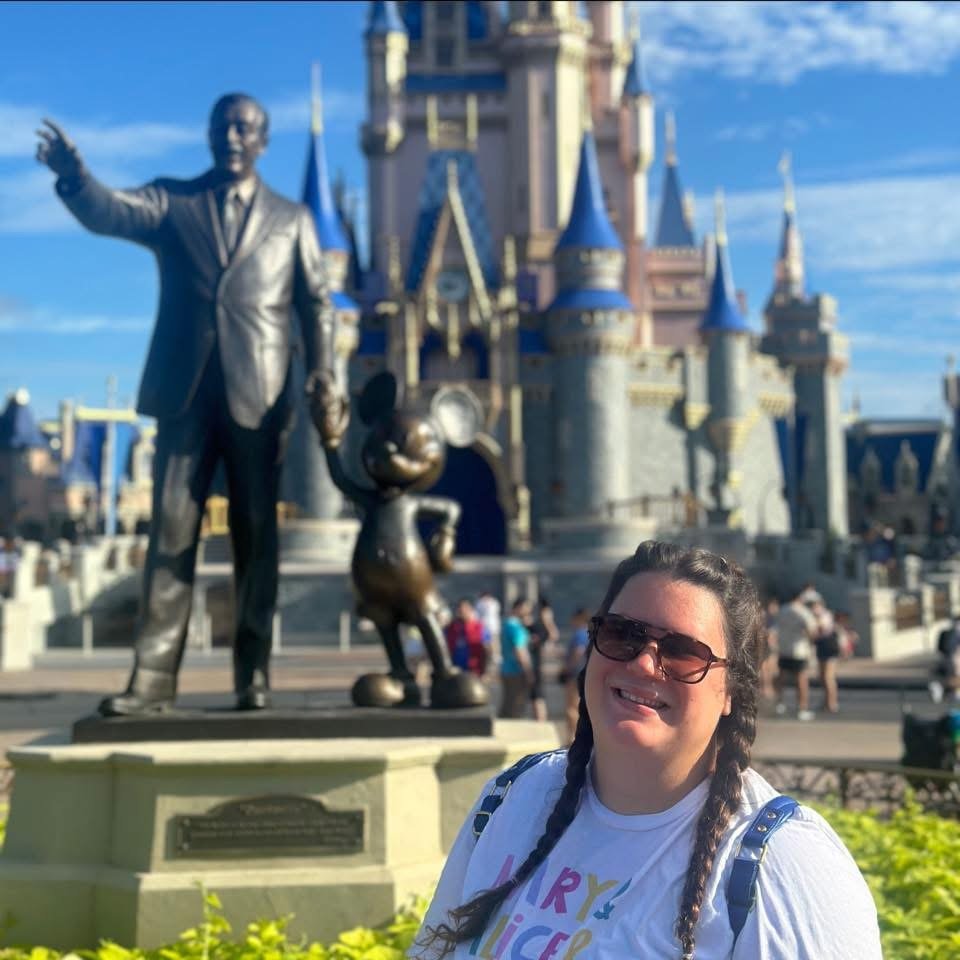 A woman with braided hair and sunglasses smiling in front of a Disney castle with a statue of Walt Disney, Mickey Mouse, and Minnie Mouse.