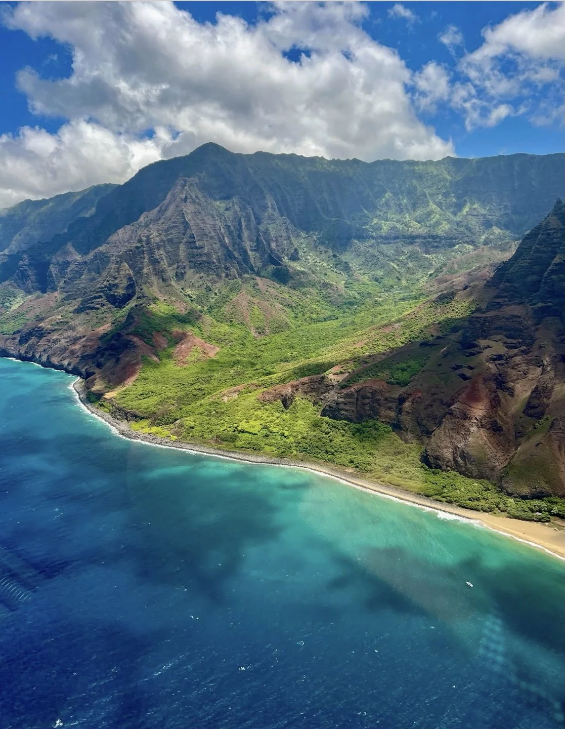 Aerial view of lush green mountains meeting a blue ocean with a sandy shoreline and partly cloudy sky.