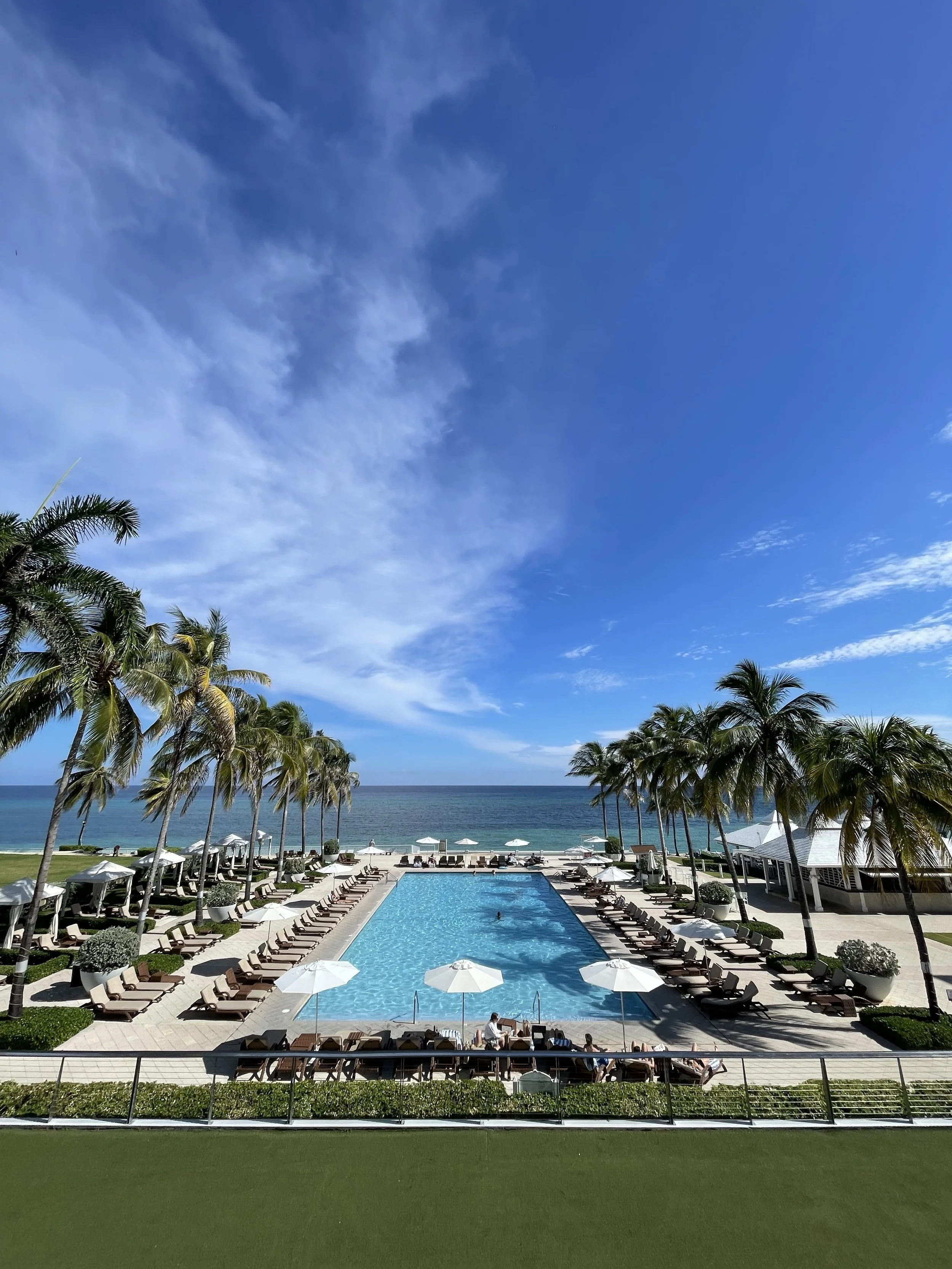 Poolside view with lounge chairs and umbrellas, surrounded by palm trees, overlooking the ocean on a sunny day.