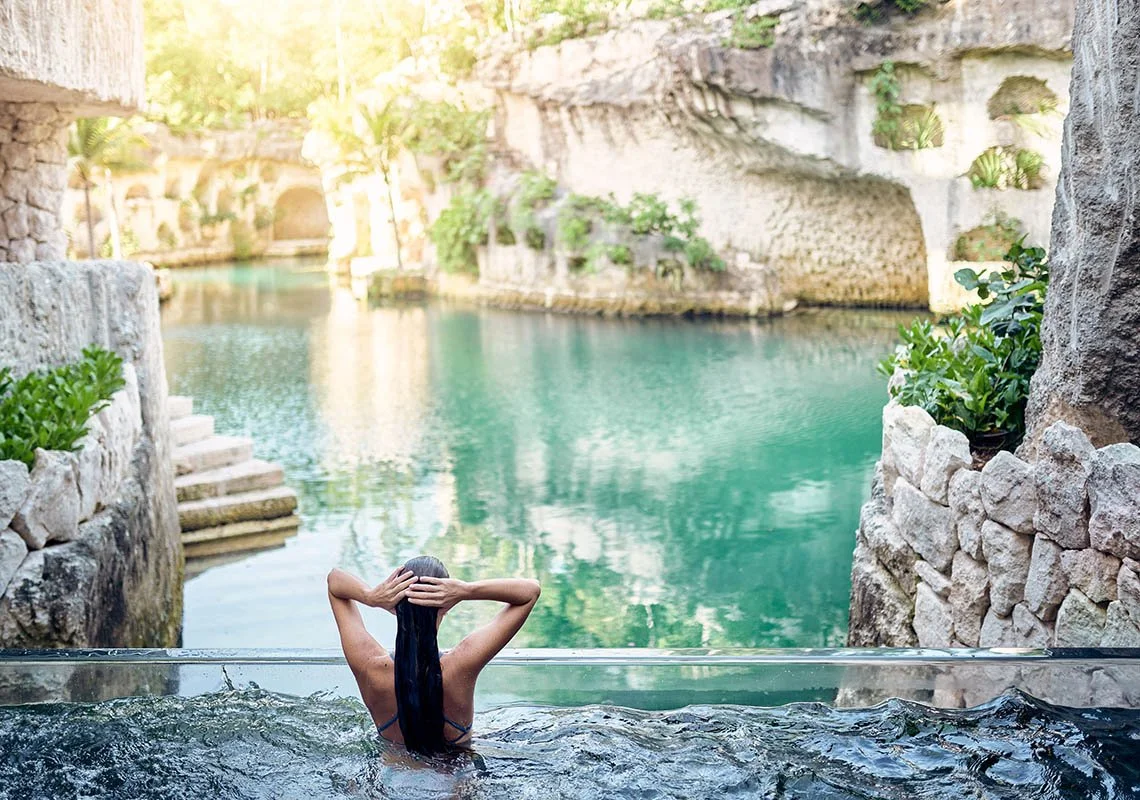 Woman with long dark hair relaxing in a natural pool surrounded by stone and greenery with calm water and a stone cave in the background.