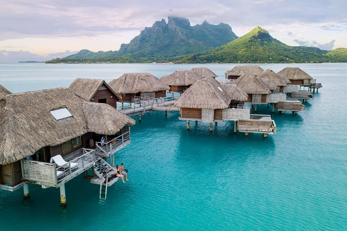 Overwater bungalows with thatched roofs in a tropical setting, with a mountain in the background and clear blue water below.
