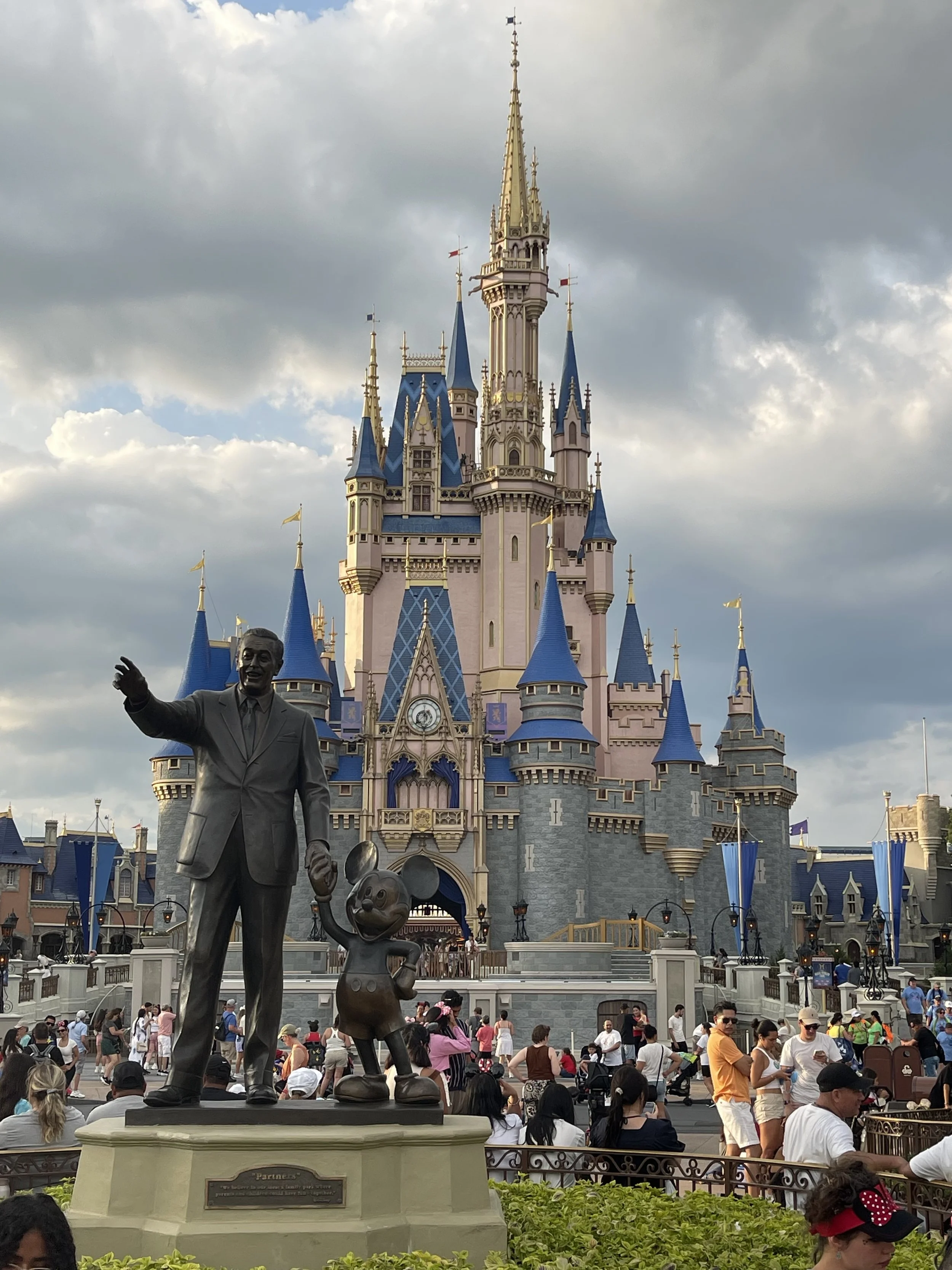 Sleeping Beauty Castle at Disneyland in the background, with a crowd of visitors in front, a bronze statue of Walt Disney and Mickey Mouse in the foreground, and cloudy skies overhead.