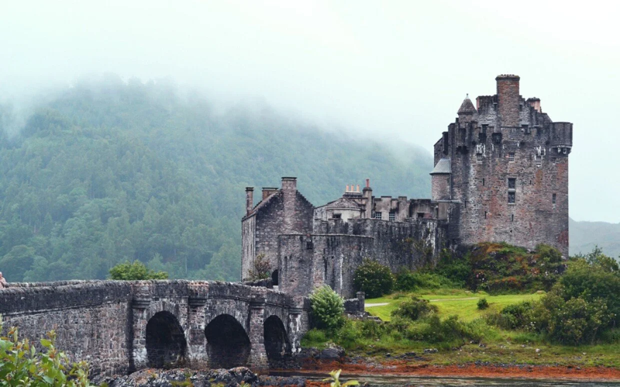 A medieval stone castle with tall towers and a surrounding wall, situated on a green landscape with a stone bridge in the foreground and misty mountains in the background.