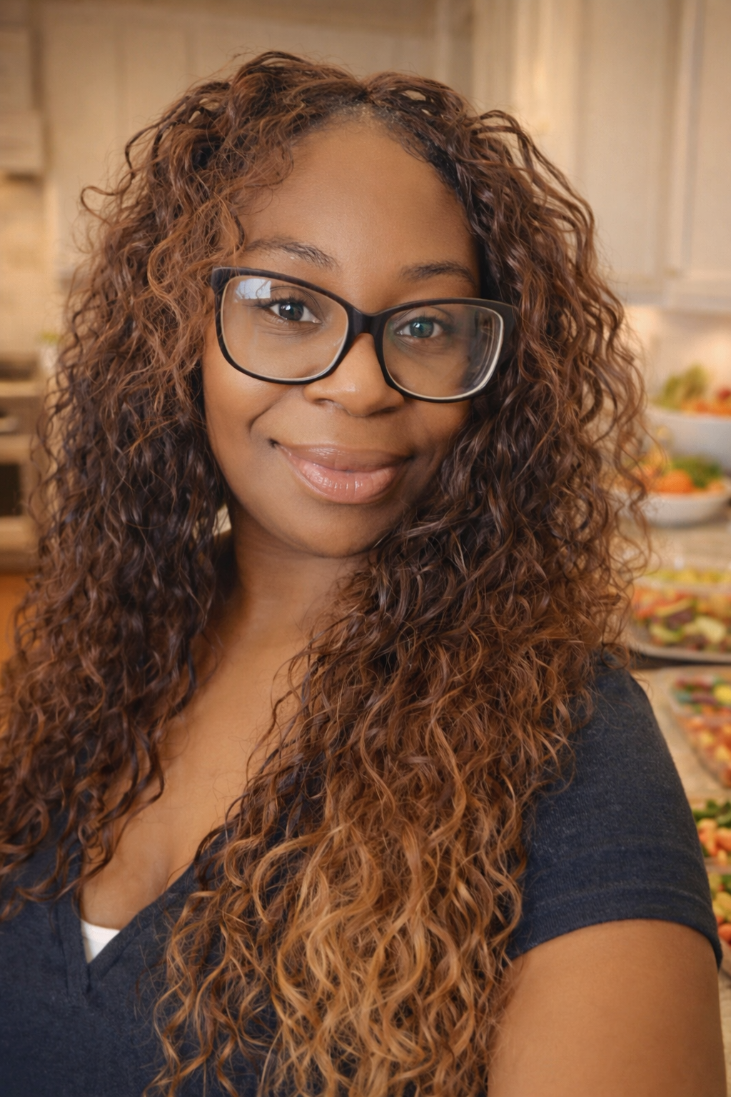 A woman with curly brown hair and glasses, smiling in a kitchen with a salad bar in the background.