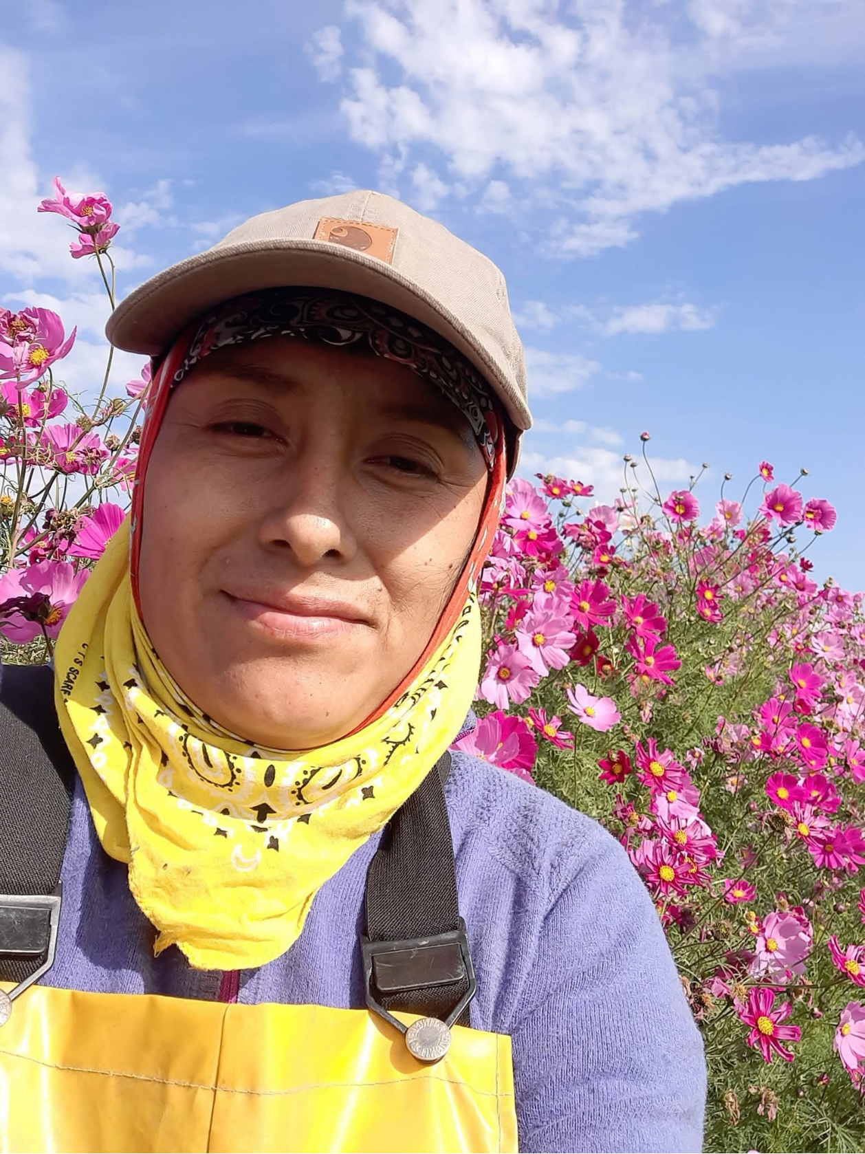A woman smiling outdoors in front of vibrant pink flowers and a blue sky with scattered clouds. She is wearing a beige cap, a black and white bandana, a yellow scarf, and a yellow apron.