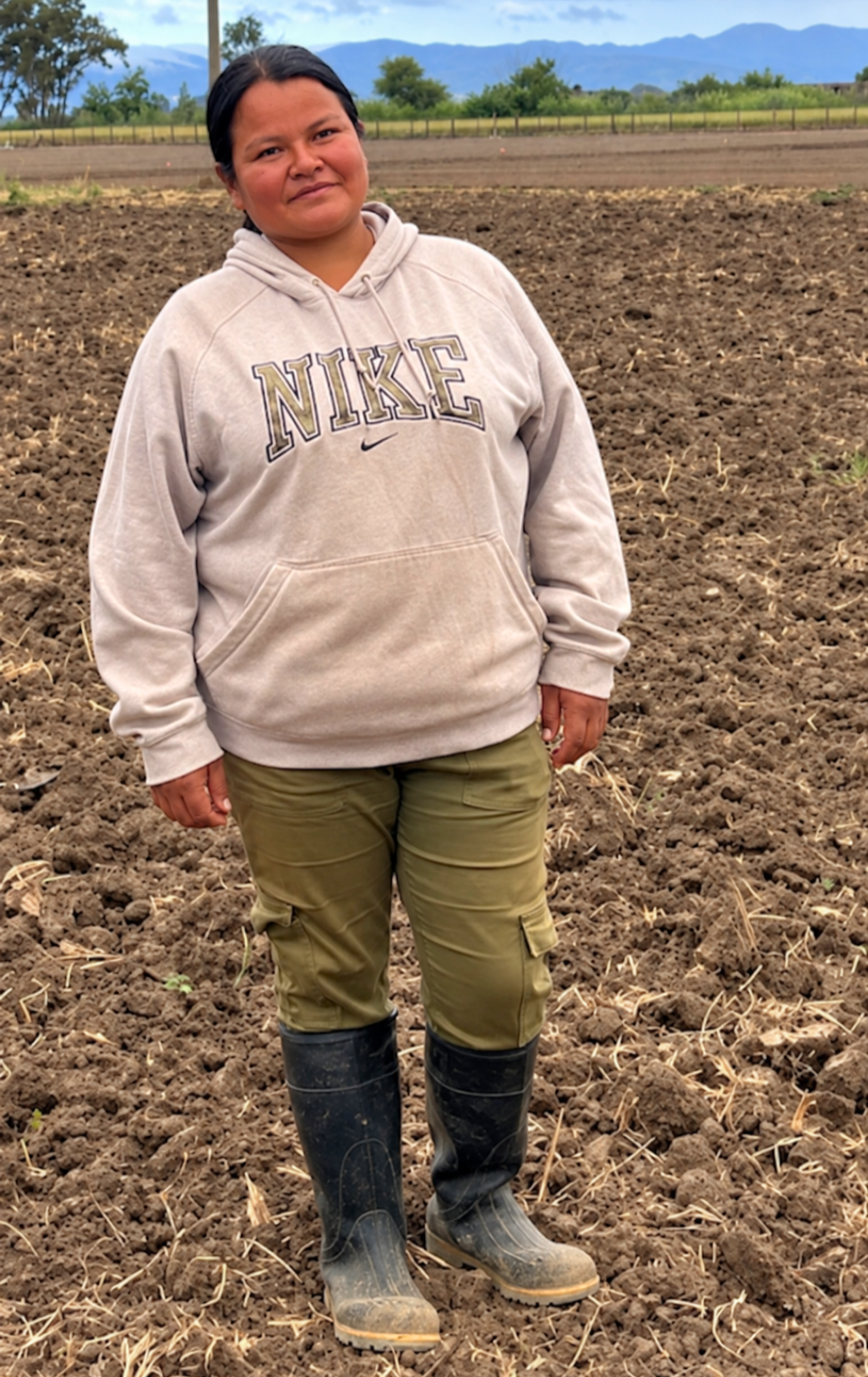 A woman standing in a plowed field with mountains in the background, wearing a beige Nike hoodie, green cargo pants, and black rubber boots.