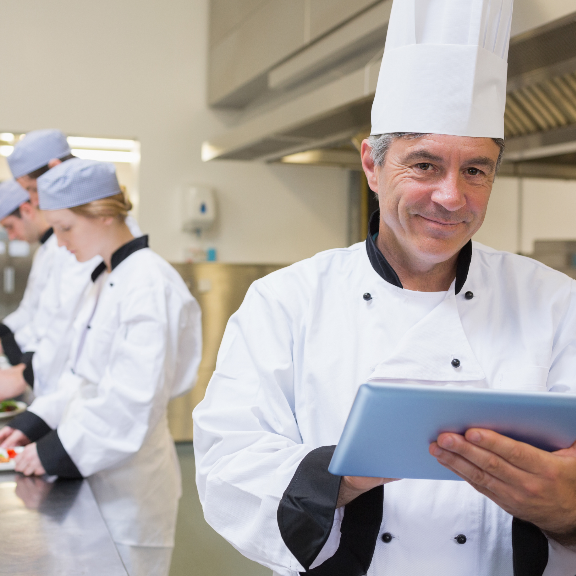Chef in uniform holding a tablet, with pastry chefs working in the background.