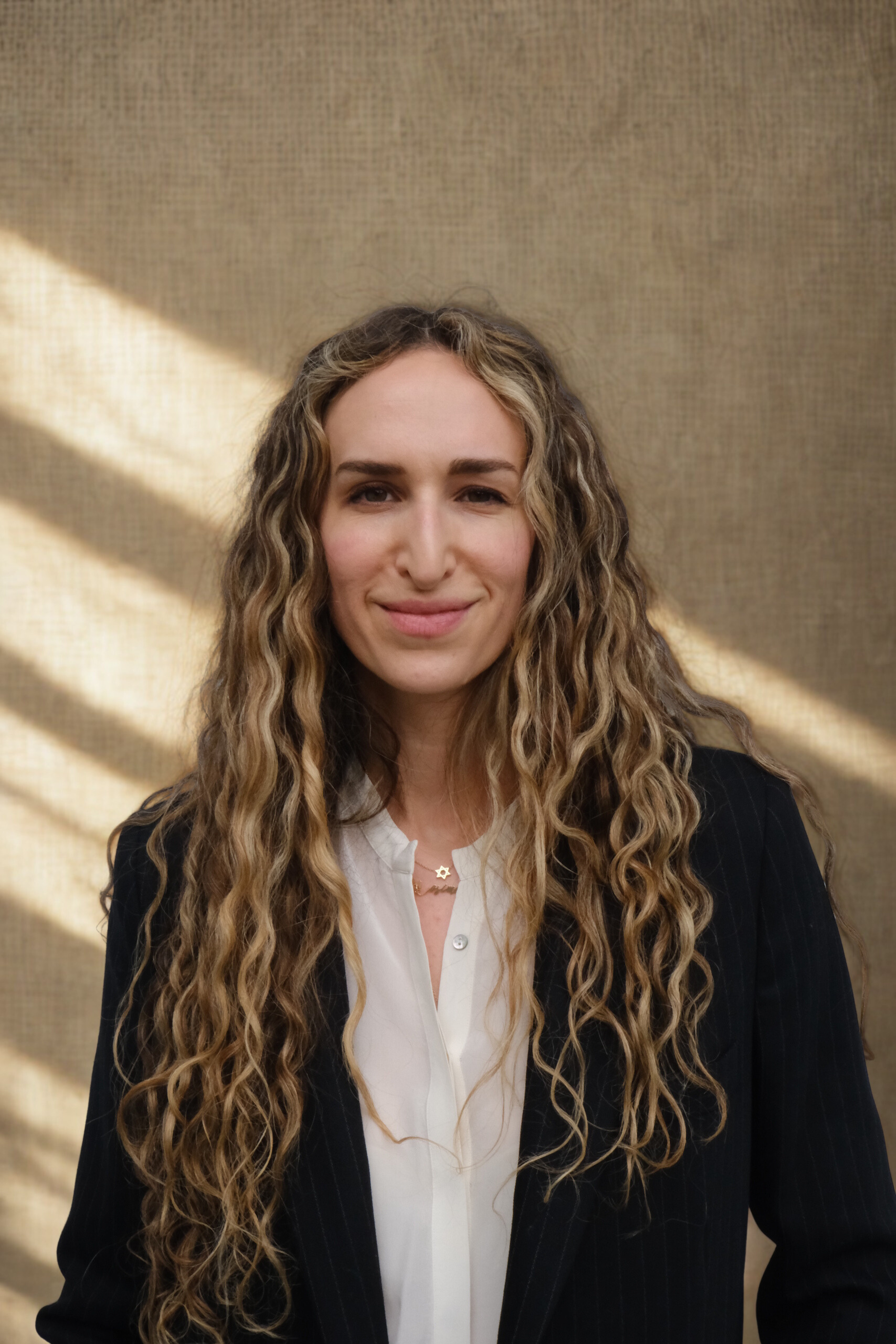 A woman with long curly blonde hair smiling, wearing a black pinstripe blazer and a white top, standing against a beige textured background with light and shadow pattern.