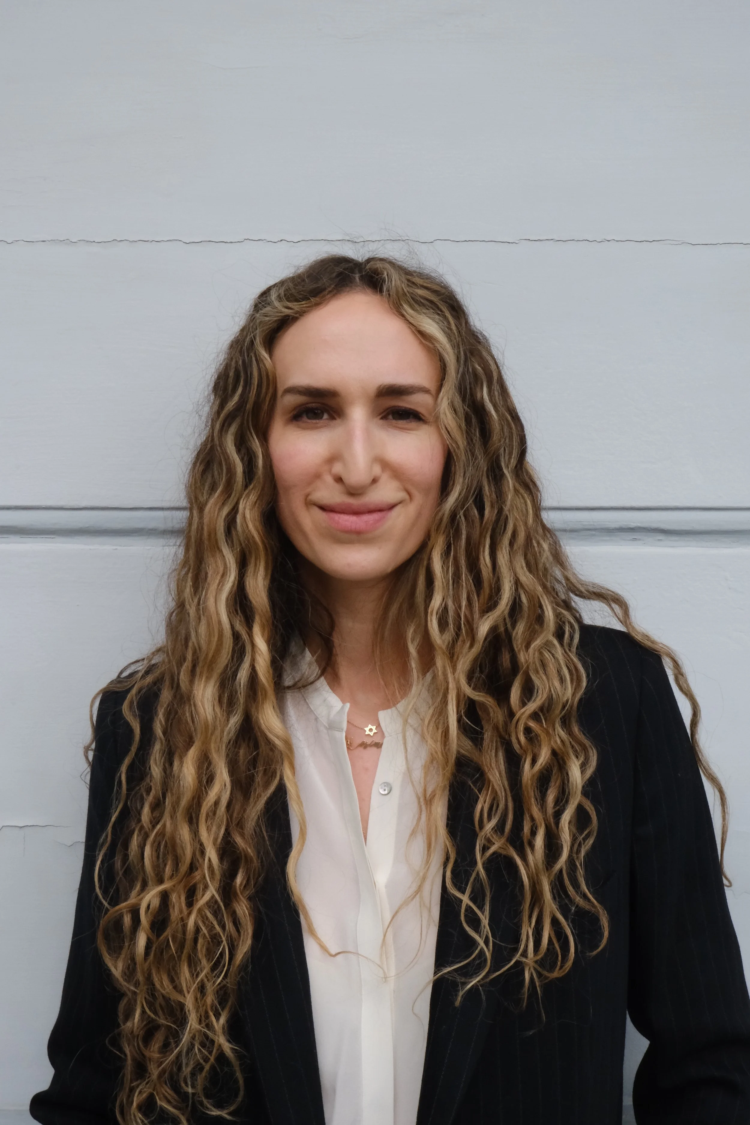 Portrait of a woman with long, curly blond hair, wearing a white blouse and a black pinstripe blazer, smiling softly in front of a plain gray wall.
