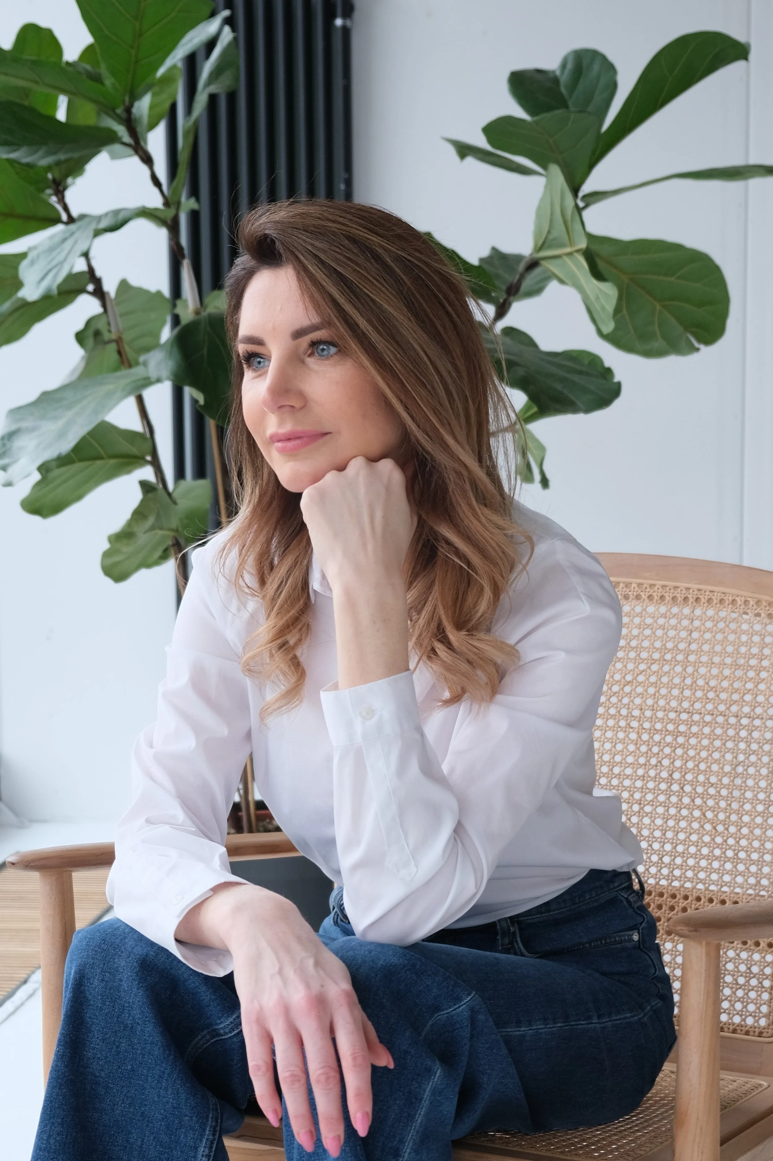 A woman with light brown hair and blue eyes, wearing a white shirt and blue jeans, sitting thoughtfully on a wooden chair with a woven back, resting her chin on her hand, in front of a large green plant with broad leaves.