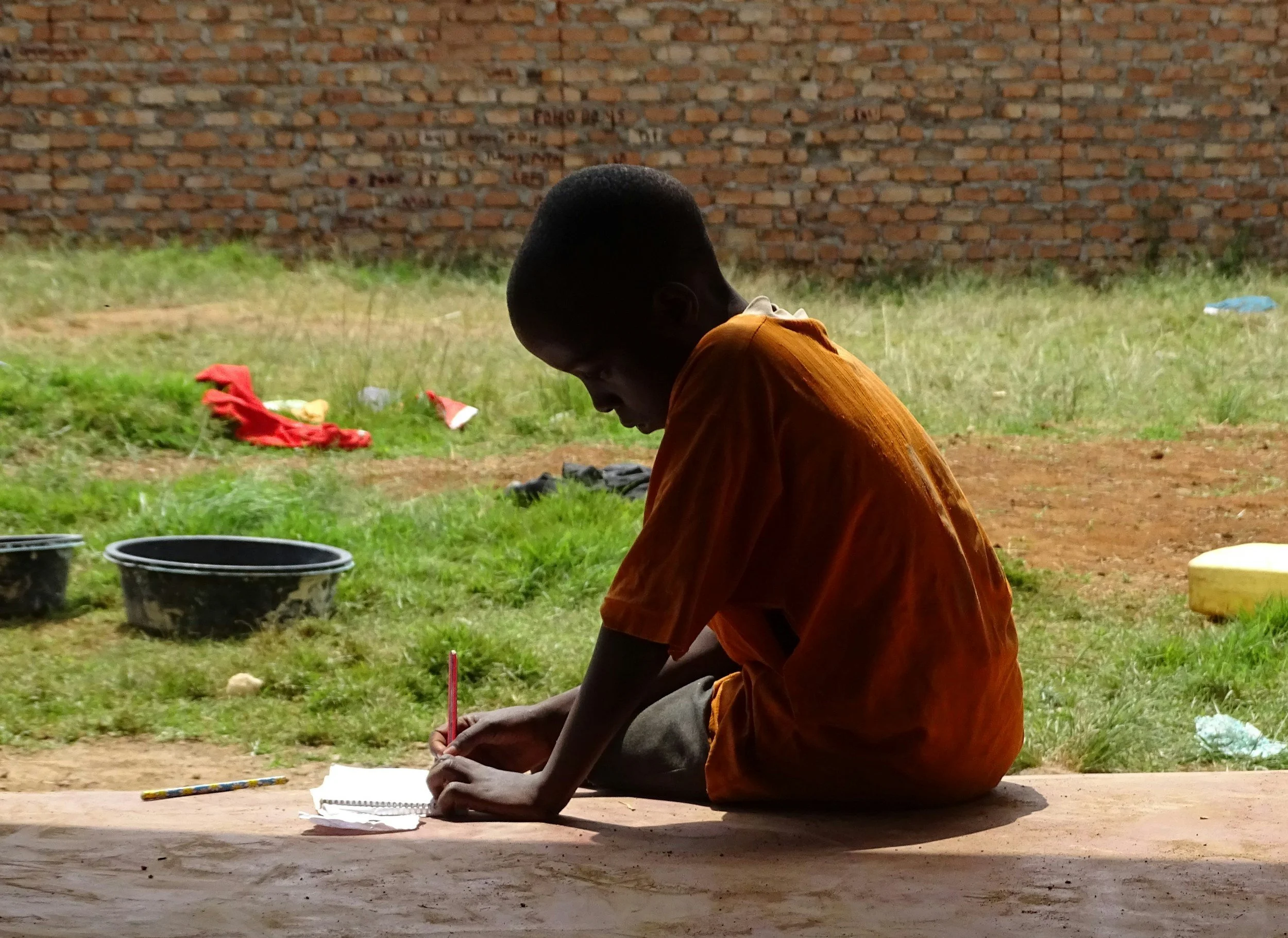 A young boy dressed in an orange shirt sitting on the ground writing or drawing on a piece of paper with a pink pencil, outdoors near a brick wall.