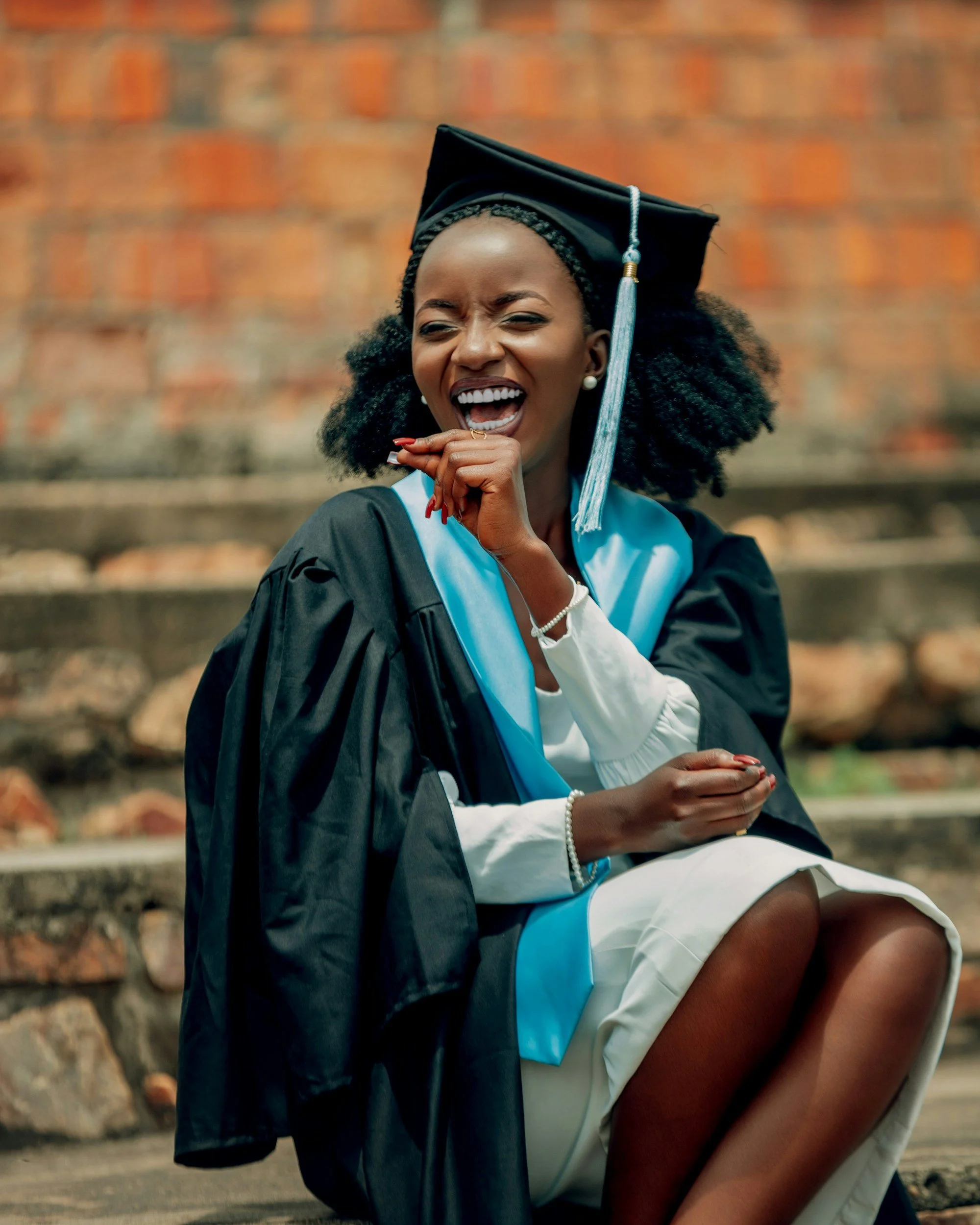 A woman in graduation attire, a cap and gown, smiling and sitting outdoors on steps with a brick wall in the background.